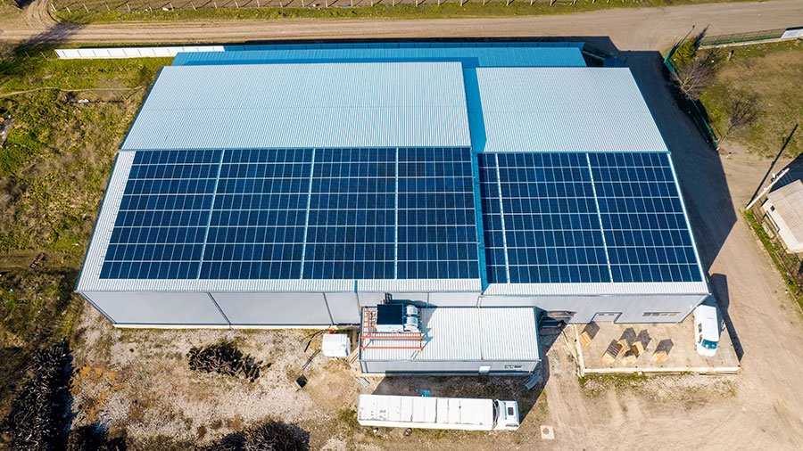 Aerial view of a large industrial building with solar panels on its roof, located near a dirt road and surrounding trees.