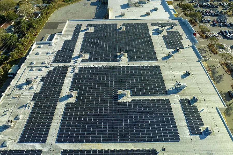 Aerial view of a building roof covered with multiple black solar panels and a parking lot filled with cars in the background.