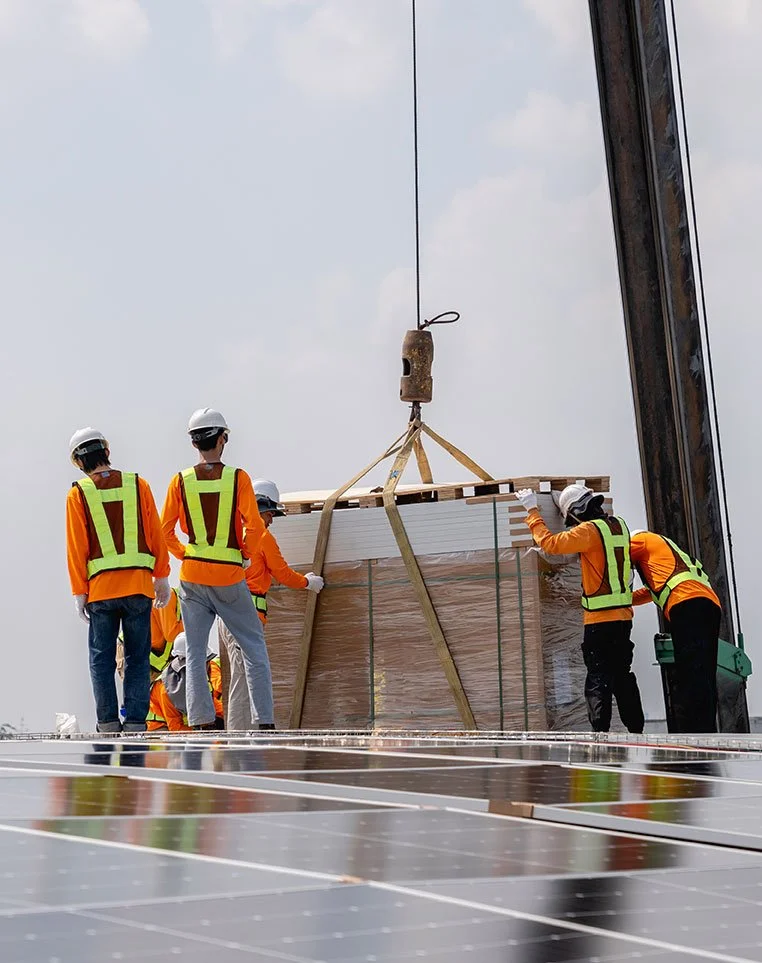 Construction workers in orange safety vests and helmets guide a crane lifting a large pallet of solar panels.