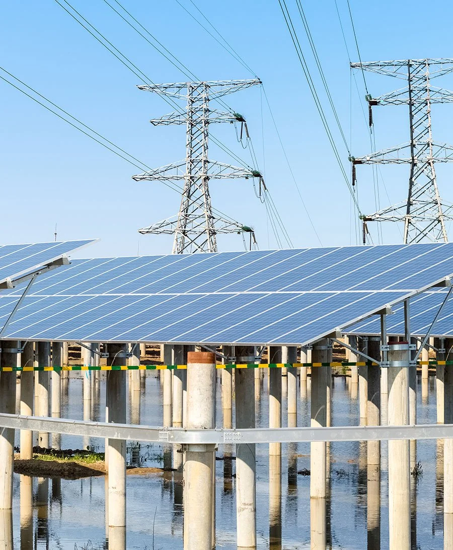 Solar panels installed on elevated supports near water, with overhead power lines and transmission towers in the background under a clear blue sky.