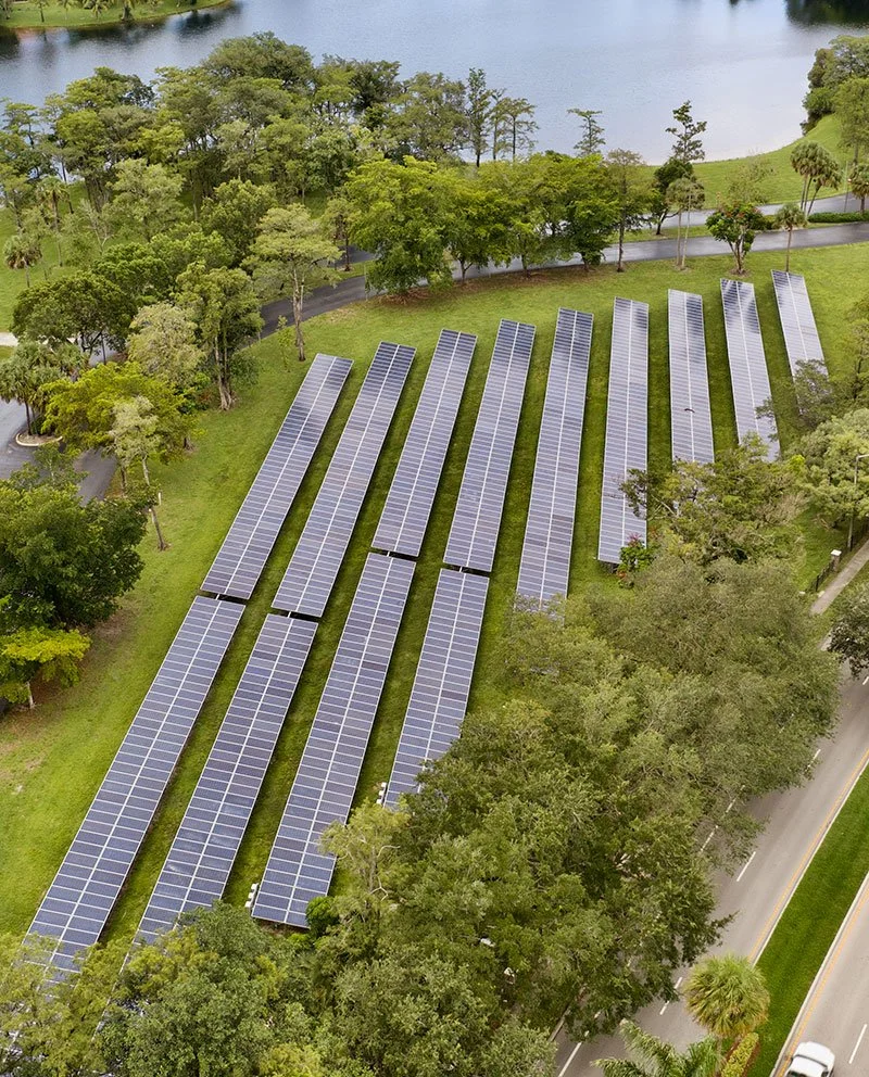An aerial view of a solar farm with multiple rows of solar panels installed on green grassy land, surrounded by trees and near a body of water.