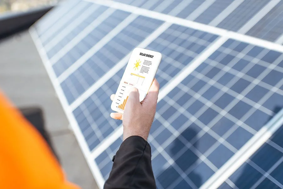 Person in an orange safety vest holding a smartphone with solar energy information displayed, standing near solar panels.