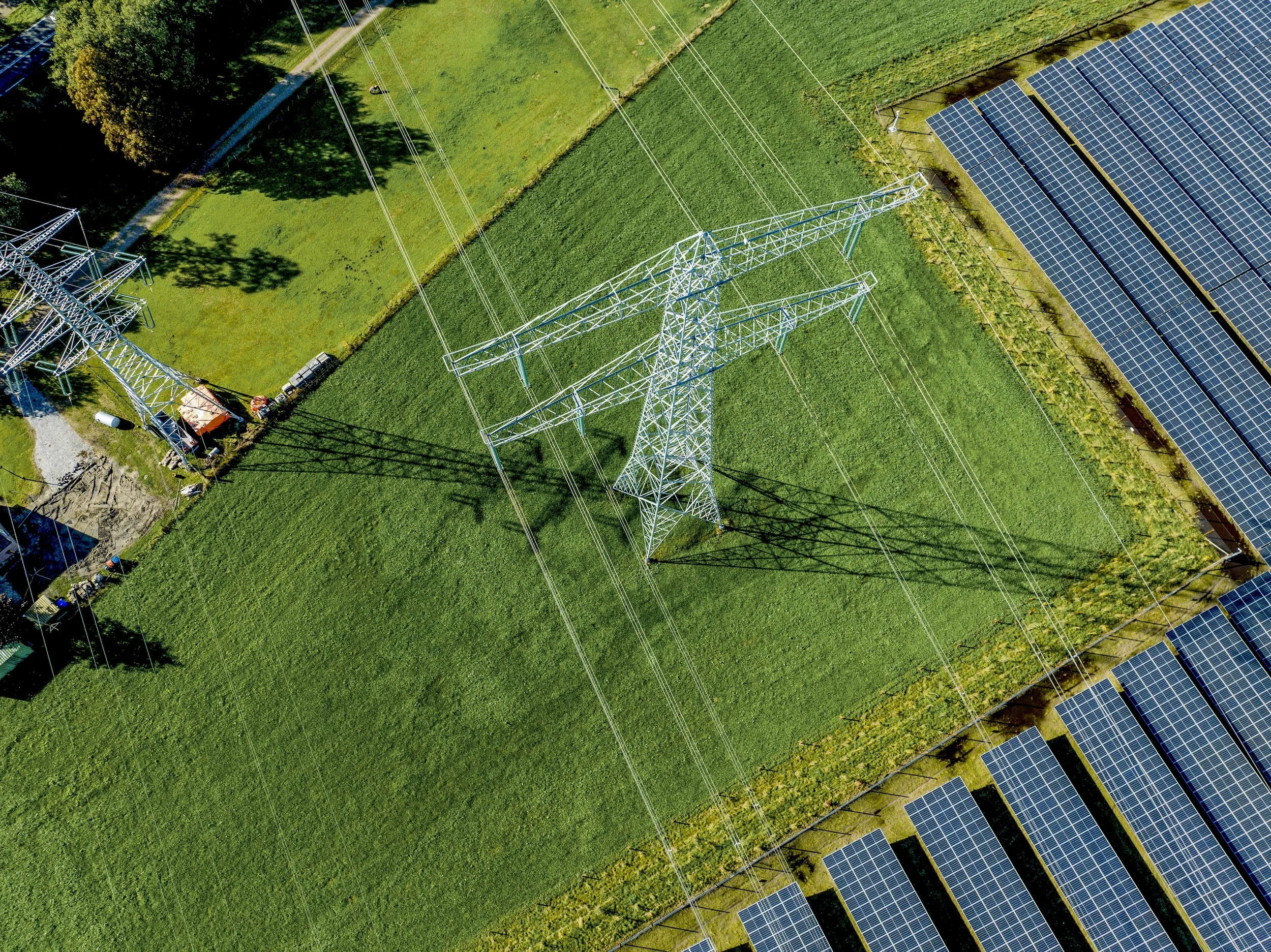 Aerial view of a green field with a transmission tower, solar panels, and power lines.