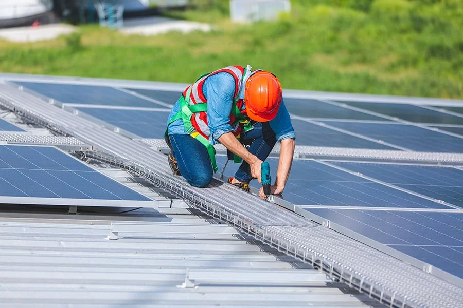 Worker installing solar panels on a metal roof, wearing a safety helmet and vest, using a power drill.