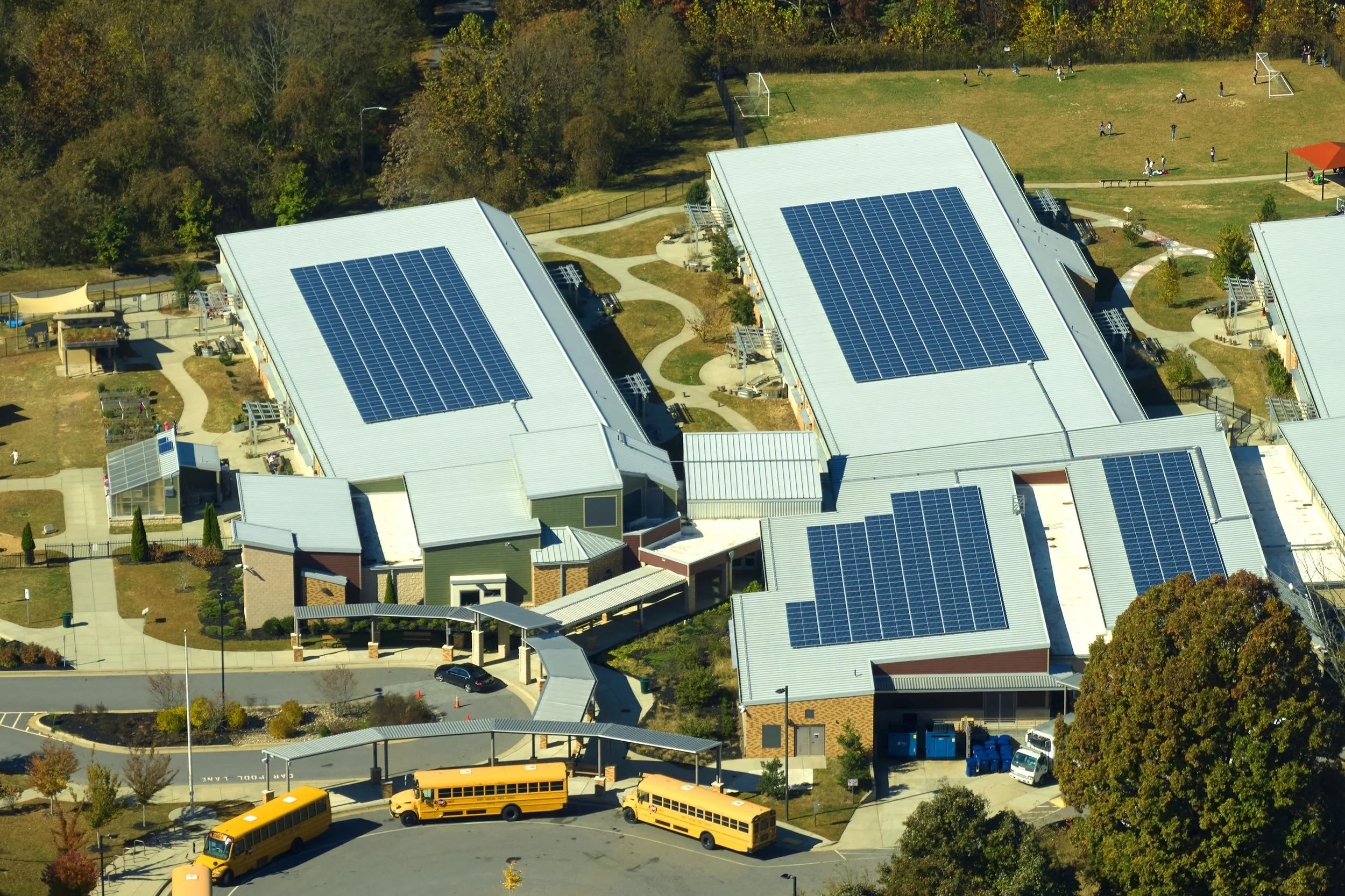 Aerial view of a school campus with multiple buildings equipped with solar panels, a football field with children playing, trees, a parking lot, and yellow school buses parked near the curb.