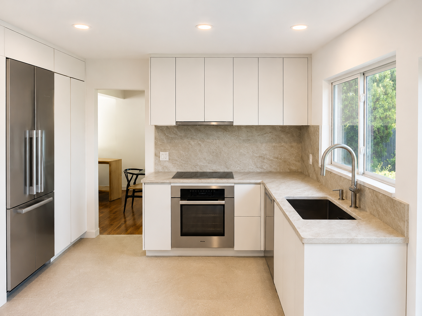 Modern kitchen with Sherwin Williams origami white cabinets, Taj Mahal counter and backsplash, stainless steel refrigerator, stainless steel sink, and view of greenery through a window.