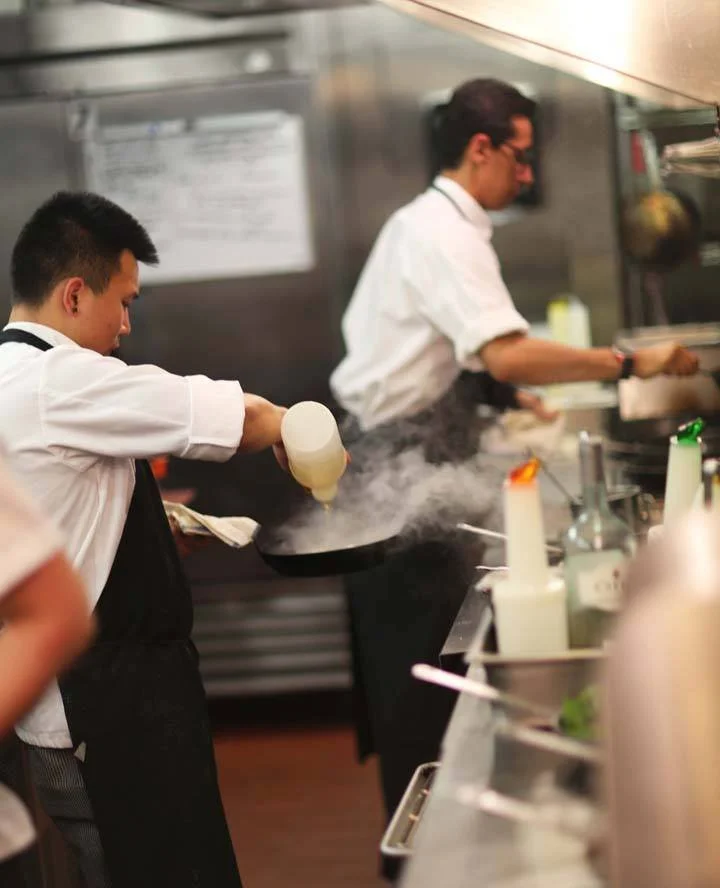 Two chefs in a professional kitchen preparing dishes, with the chef in the foreground pouring sauce onto a plate and the chef in the background cooking on a stove.