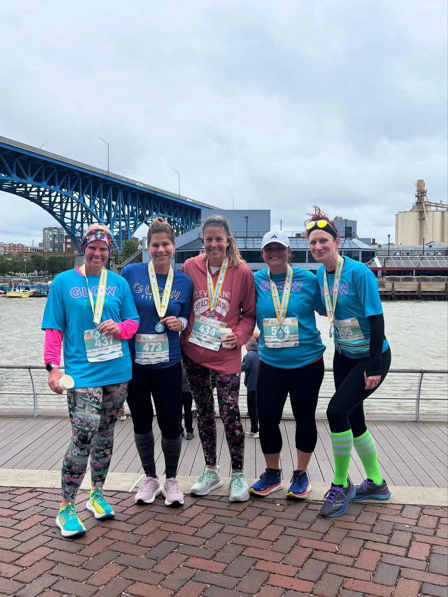 Group of five women standing on brick walkway with medals around their necks, near river, with bridge and industrial buildings in background, after running race.