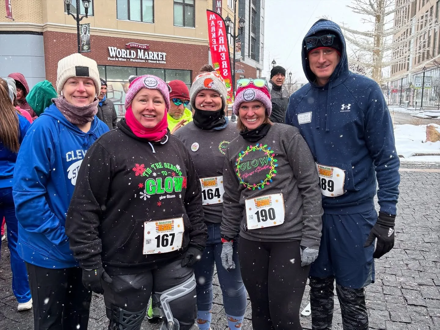 Group of five smiling people in running attire and winter gear, standing outside in snowy weather, participating in a race event, with a city street and store signs in the background.