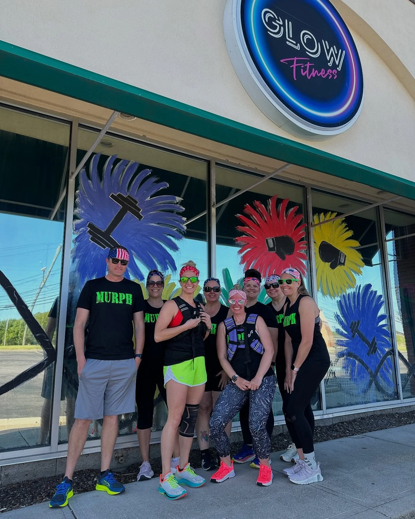 A group of seven people wearing athletic clothes and sunglasses standing outside a fitness gym called Glow Fitness, with colorful feather and boxing glove artwork on the windows behind them.