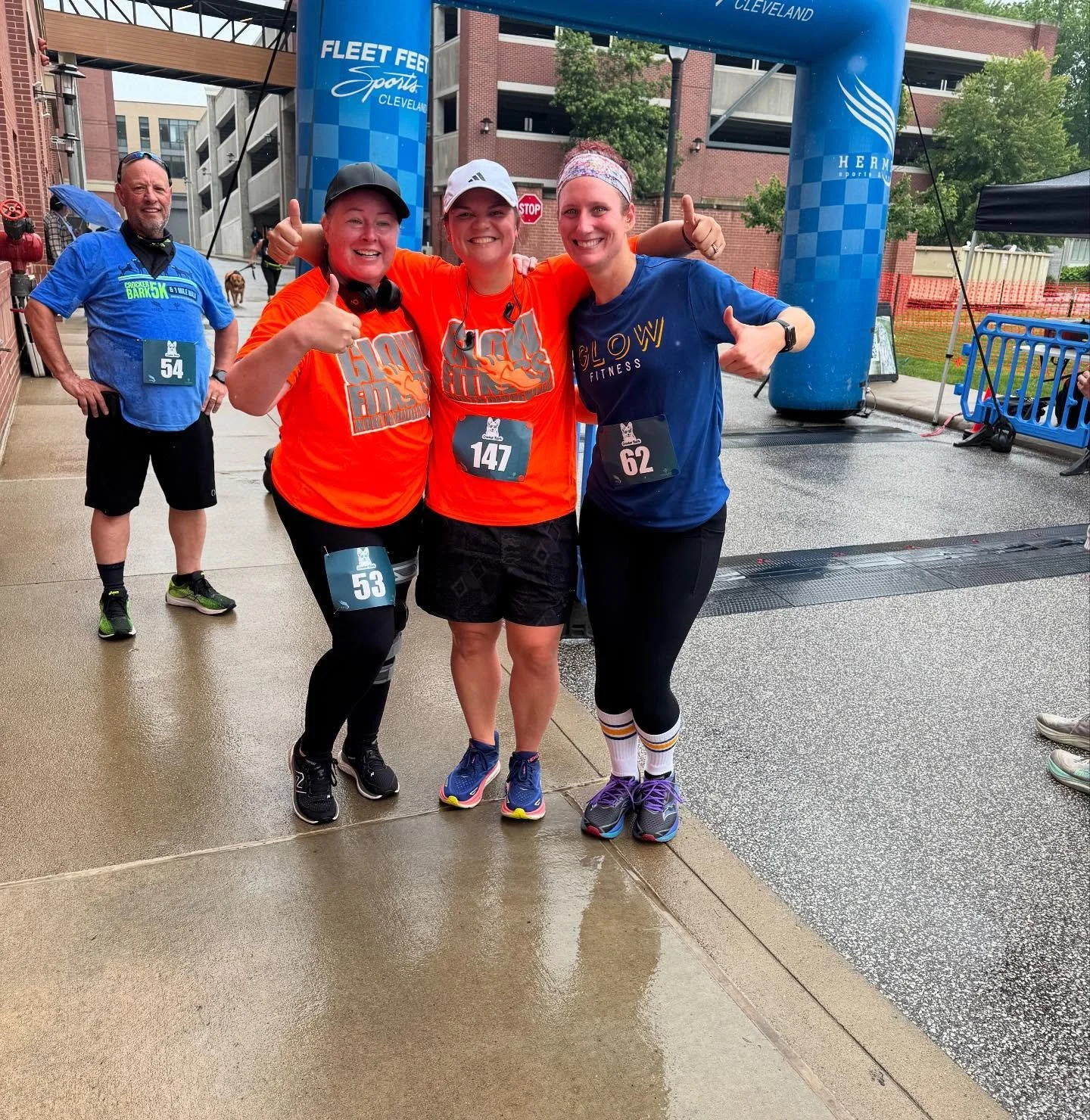 Three women and one man at a finish line for a running event, smiling and giving thumbs up, wearing athletic clothing and numbered bibs. The background features a blue inflatable arch and a wet pavement with a building and trees.