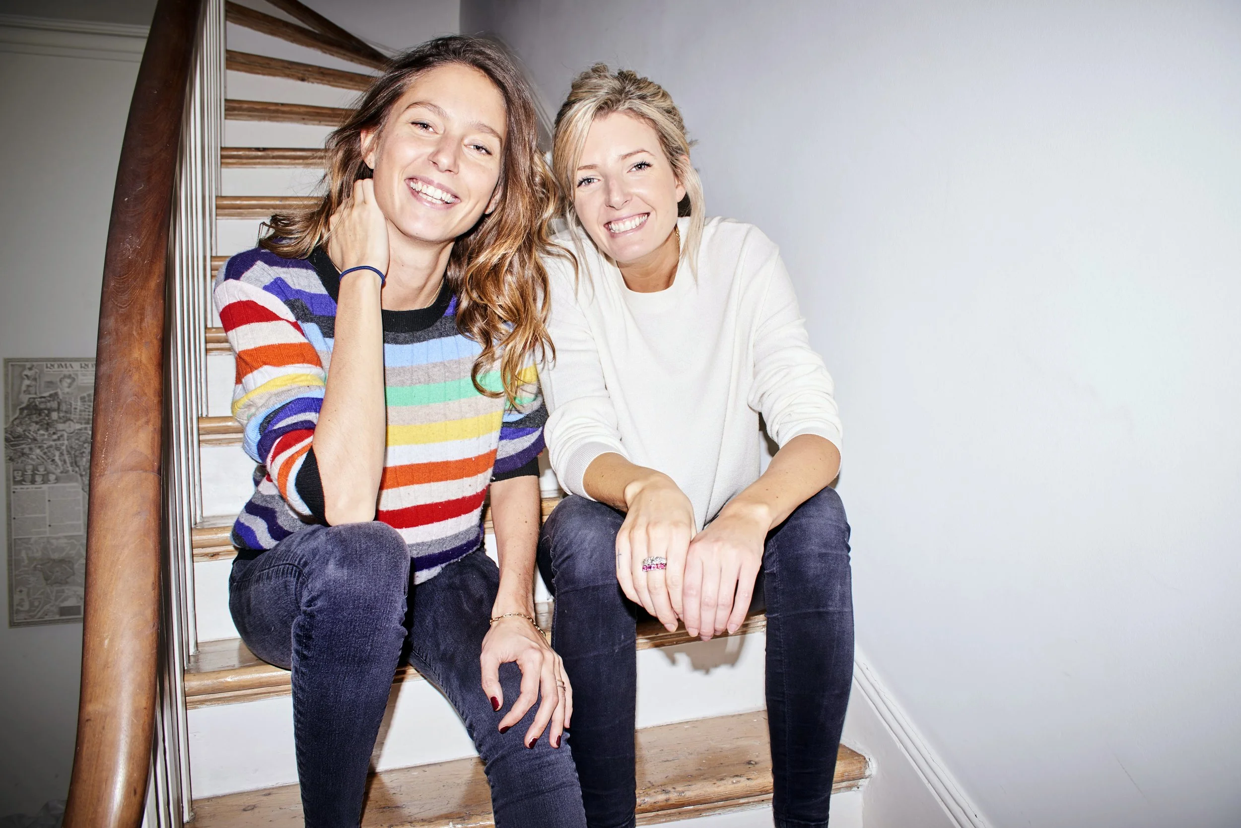 Two women sitting on a staircase, smiling at the camera. One woman has long, wavy hair and is wearing a multicolored striped sweater. The other woman has blonde hair and is wearing a cream-colored top. The staircase is wooden with a simple white wall background.