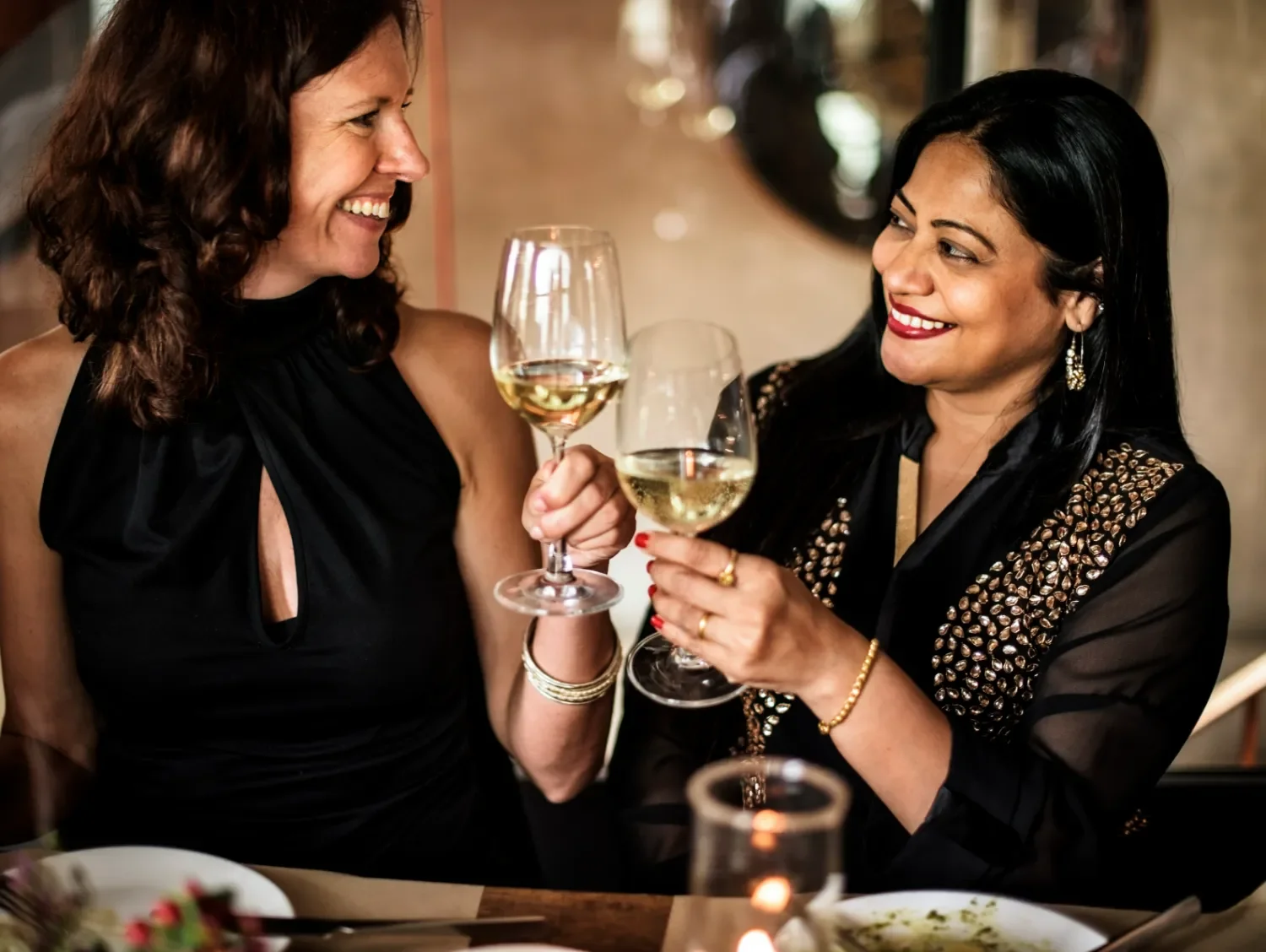 Two women dressed in chic black outfits, toasting wine glasses with white wine