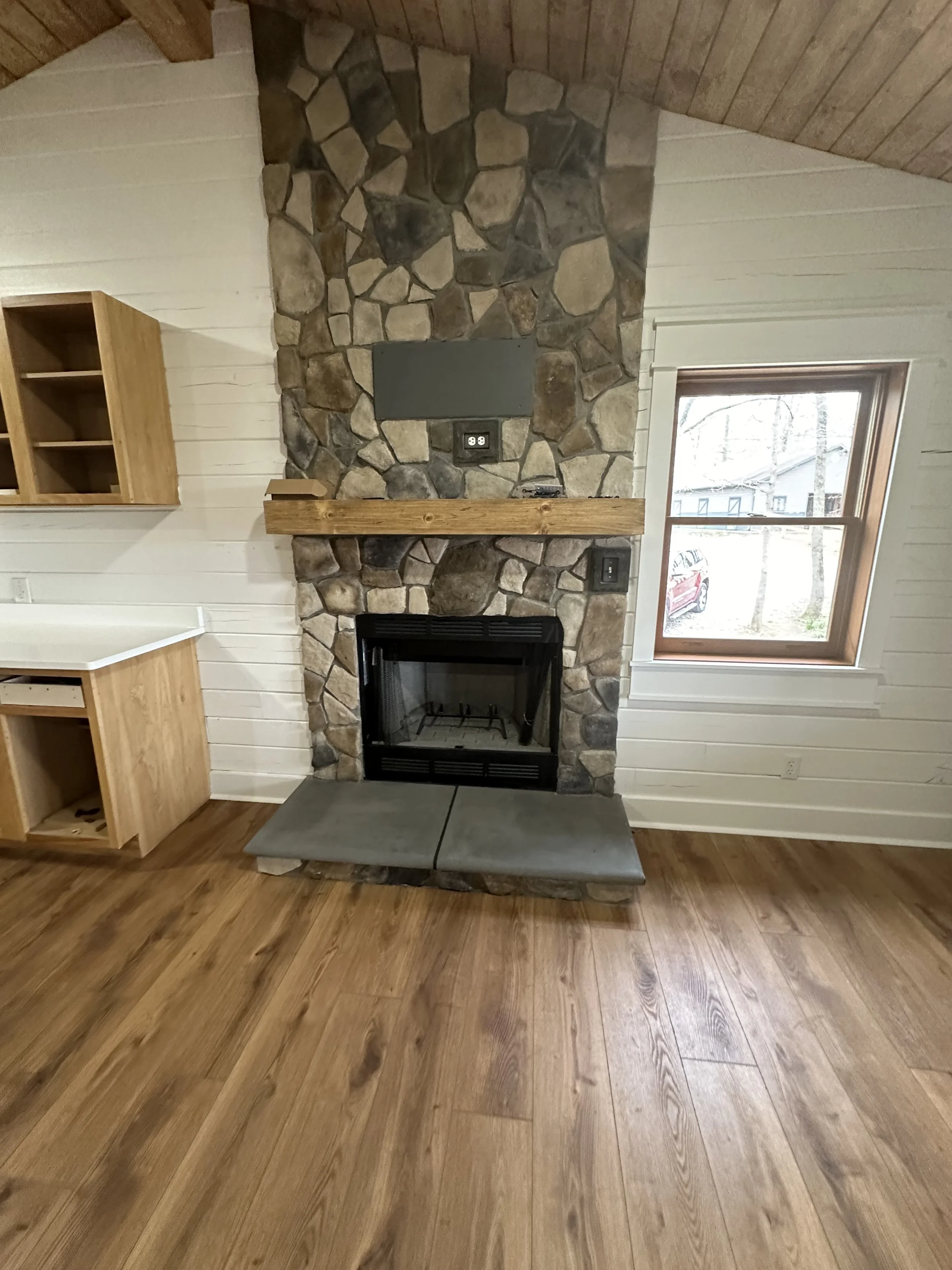 Living room with stone fireplace, wooden mantel, and a window showing a rural scene outside
