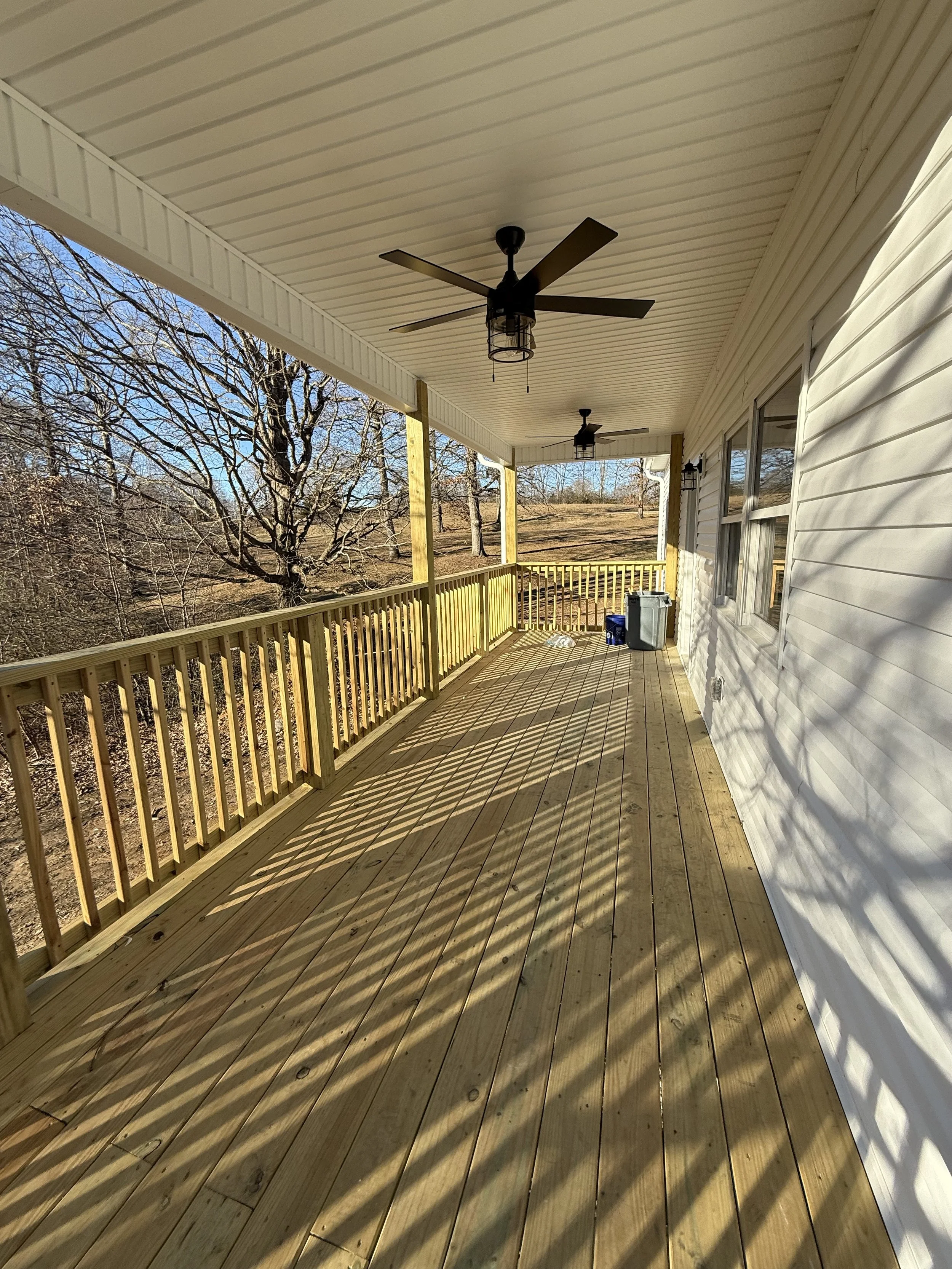 A spacious outdoor wooden porch with ceiling fans, overlooking a yard with leafless trees, enclosed with a yellow wooden railing, during the daytime.