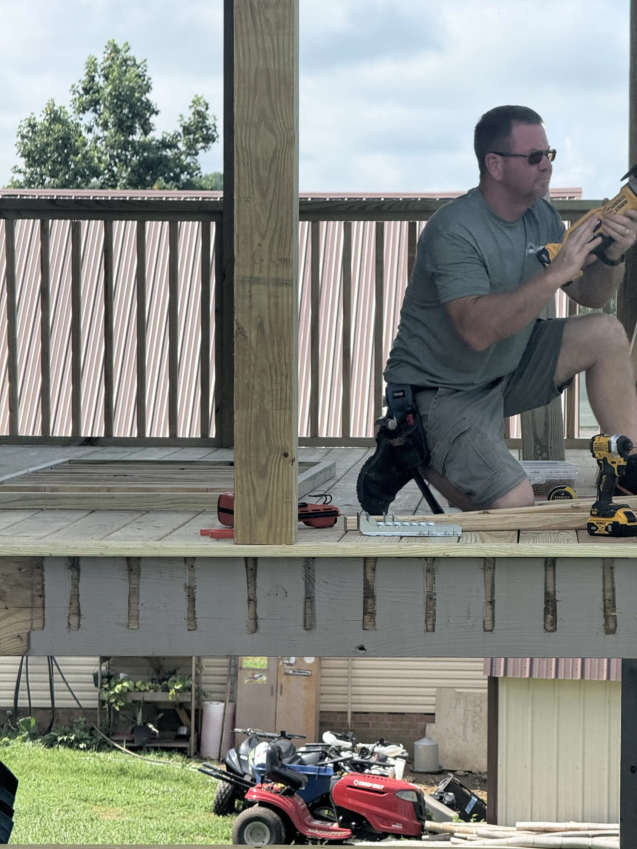 Man kneeling on a wooden deck, using a power drill, surrounded by tools, with a putty knife nearby. The deck is under construction, with a wooden railing, and outdoor equipment and a toy riding lawn mower are visible below.