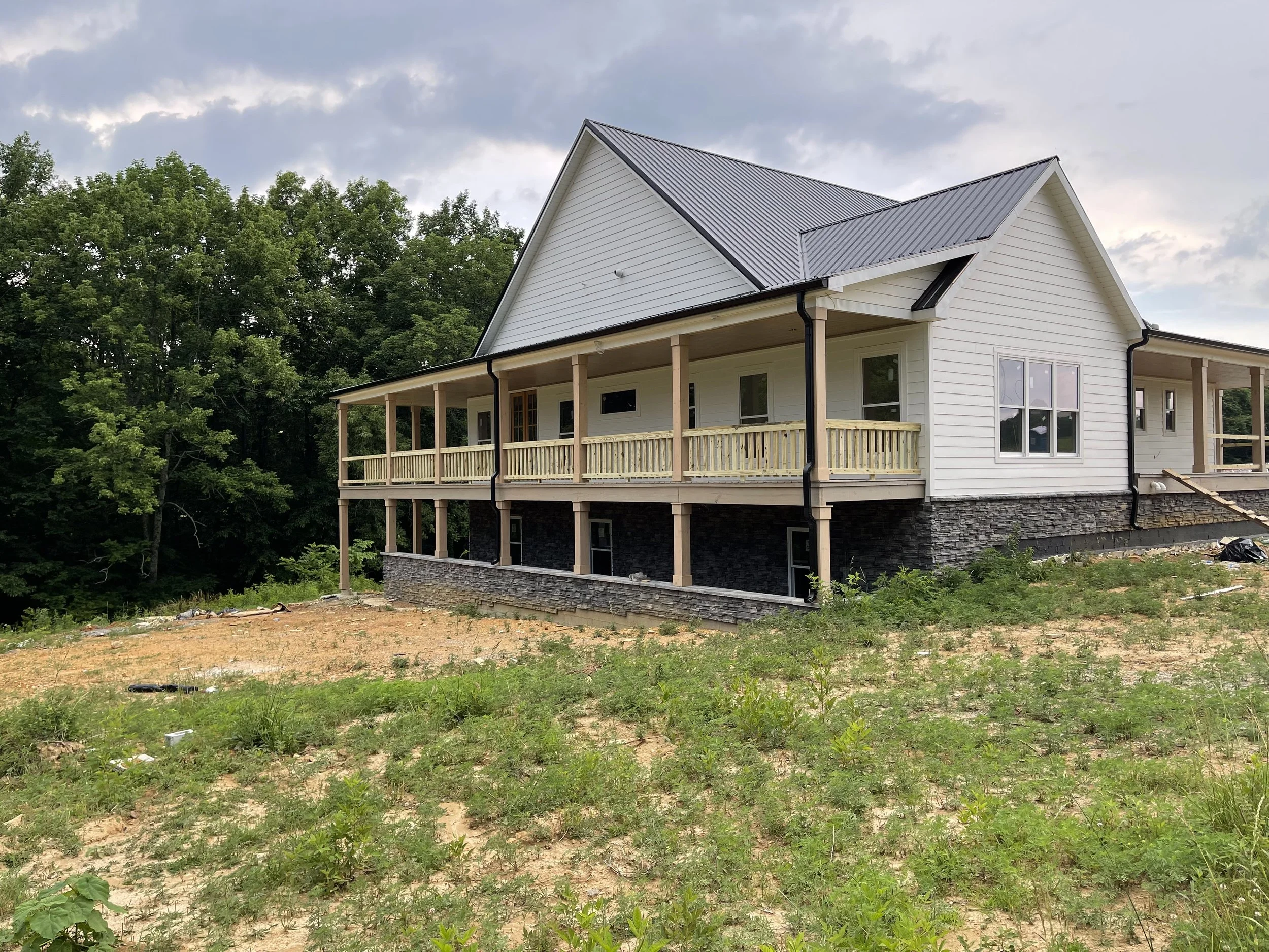 Newly built house with white siding, black roof, and a large front porch overlooking a grassy yard with patches of dirt, surrounded by trees.