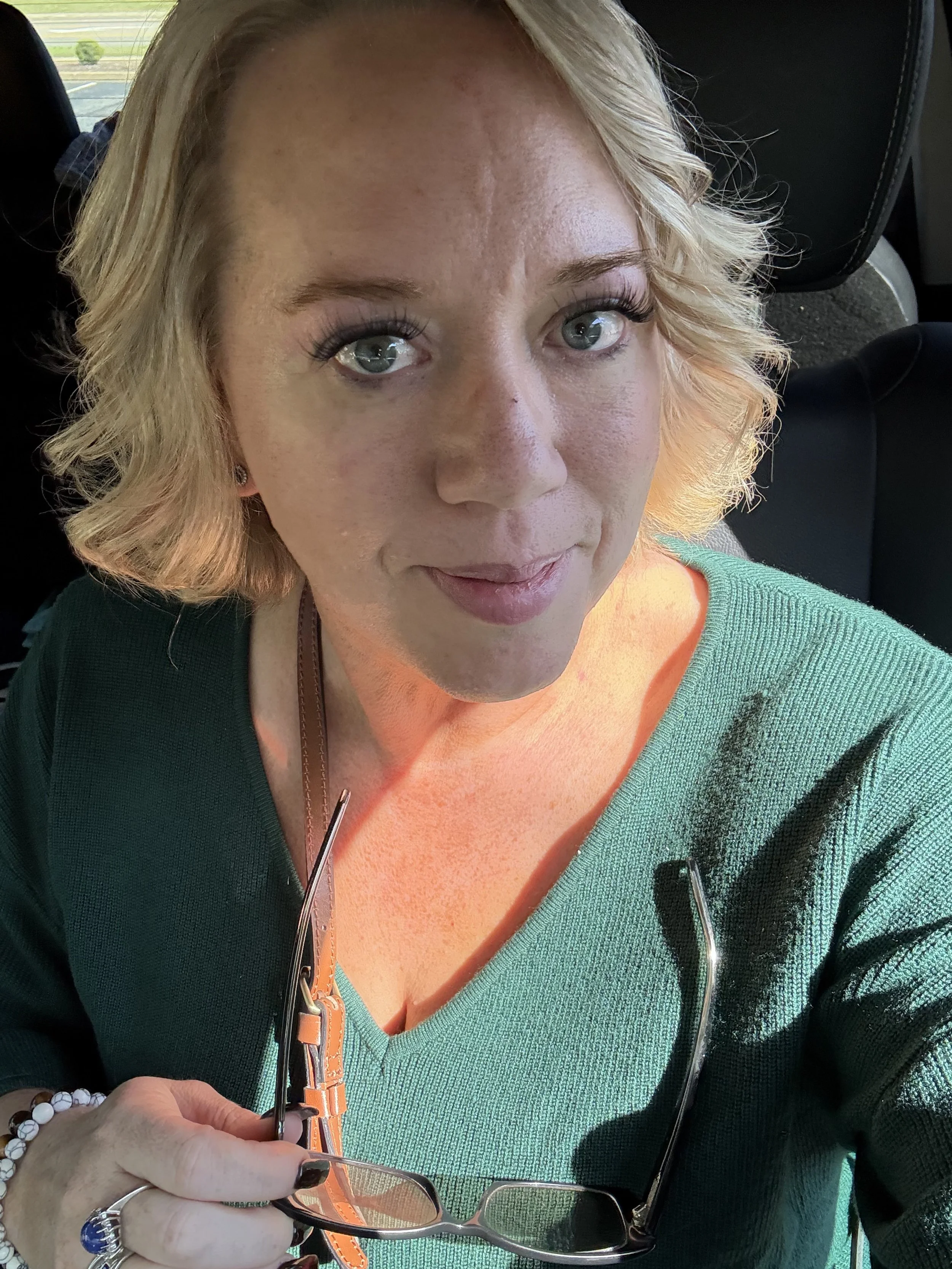Close-up of a woman with blonde hair and blue eyes, wearing a green top, sitting in a vehicle, holding glasses with a orange strap.
