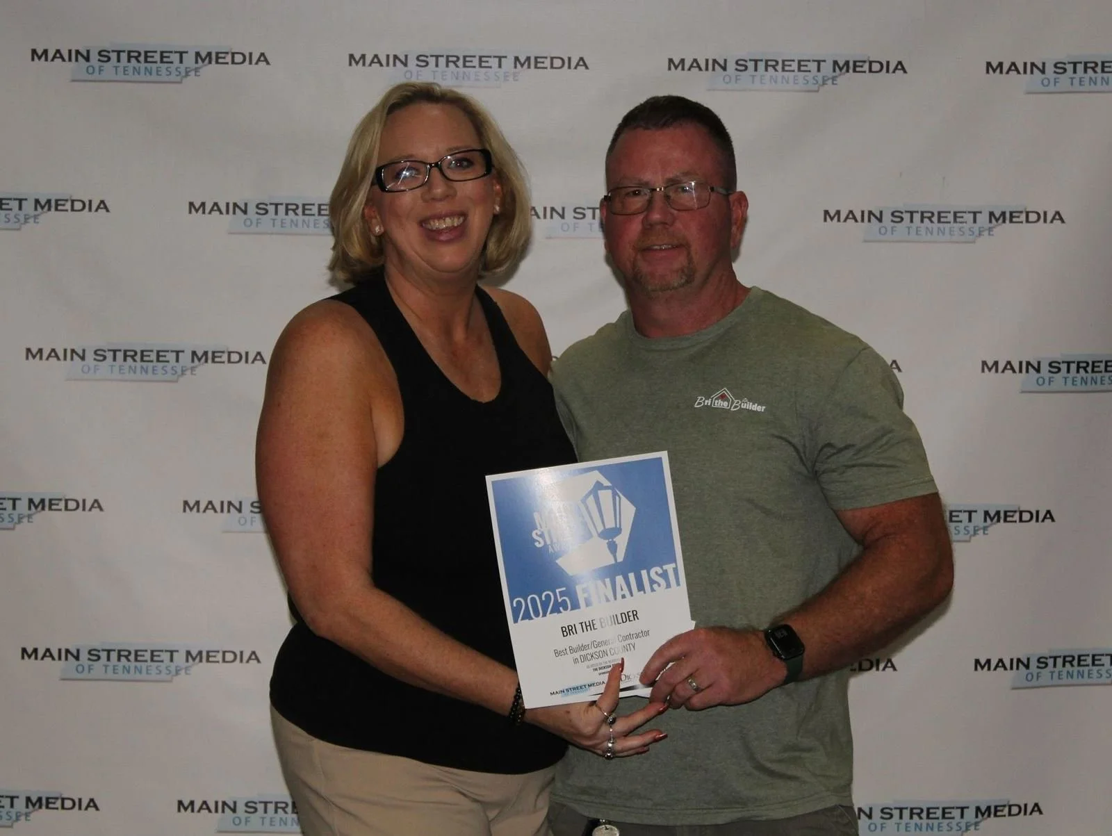 Two people, a woman with glasses and a man with glasses and a beard, holding a award plaque that says 'Finalist 2025 Best Builder General Contractor in Dickson County,' standing in front of a backdrop with the 'Main Street Media of Tennessee' logo.