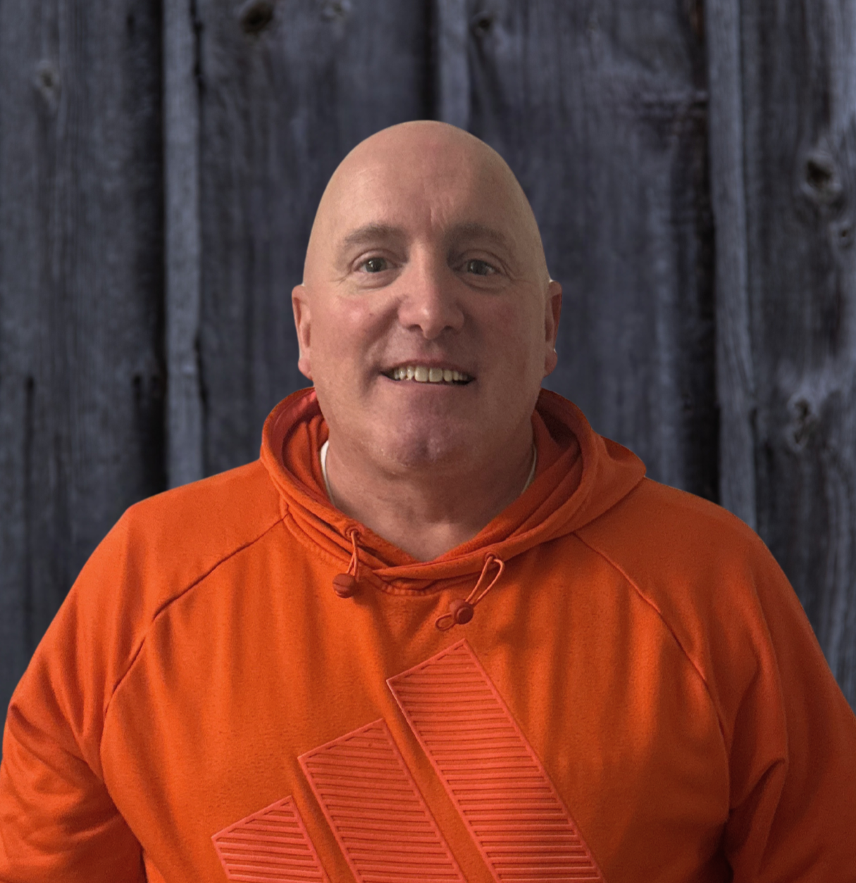 A man with a shaved head smiling, wearing an orange hoodie with graphics, standing in front of a textured wooden background.