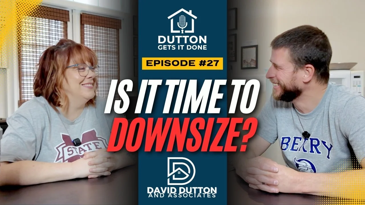 Two people sitting at a table smiling and talking in a room with framed pictures on the wall. The woman has red hair and glasses, the man has dark hair and a beard. There is a logo for 'Dutton Gets It Done' and text asking 'Is it time to downsize?'