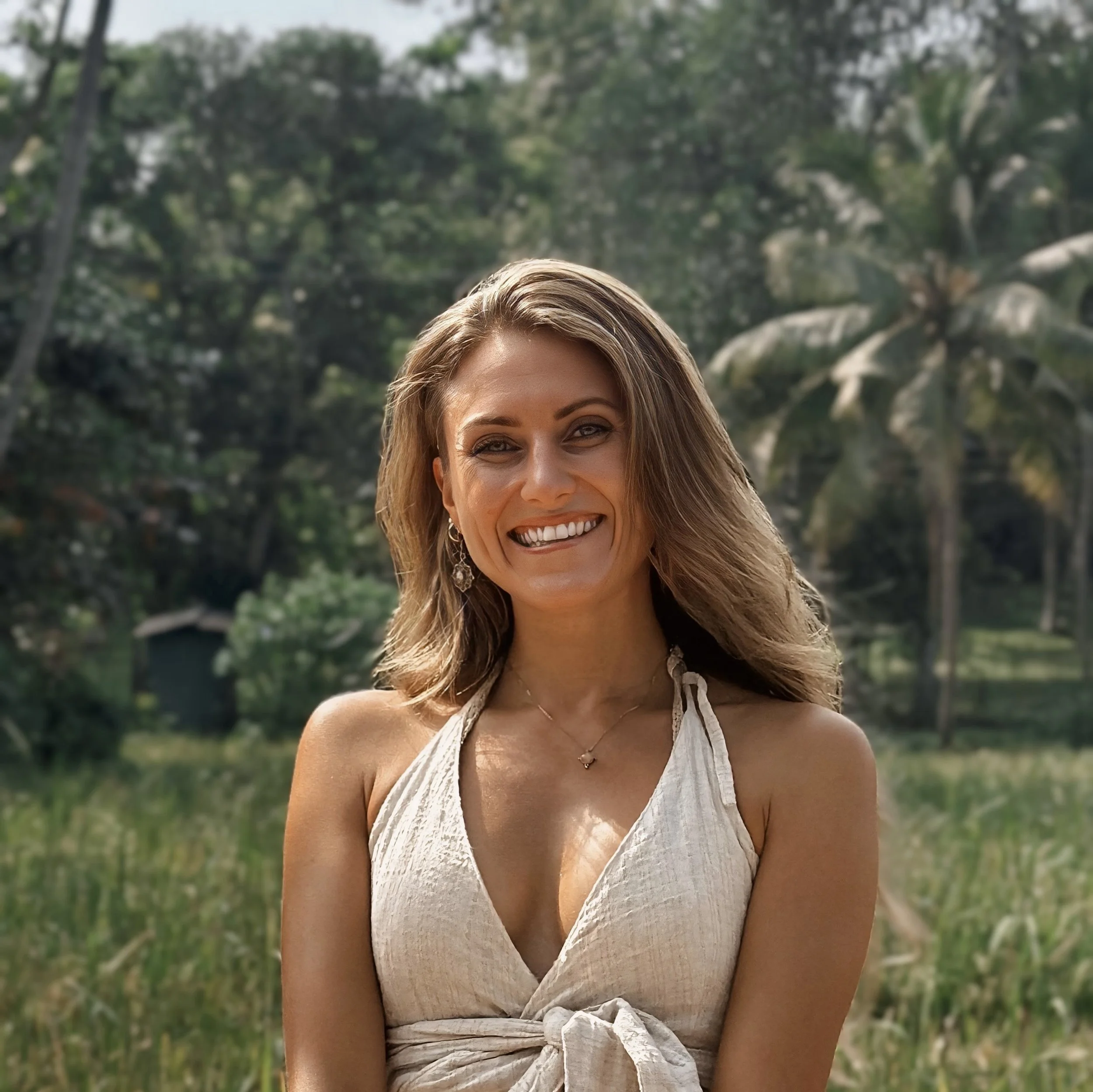 Frau sitzt lächelnd an einem Ort umgeben von Palmen. 