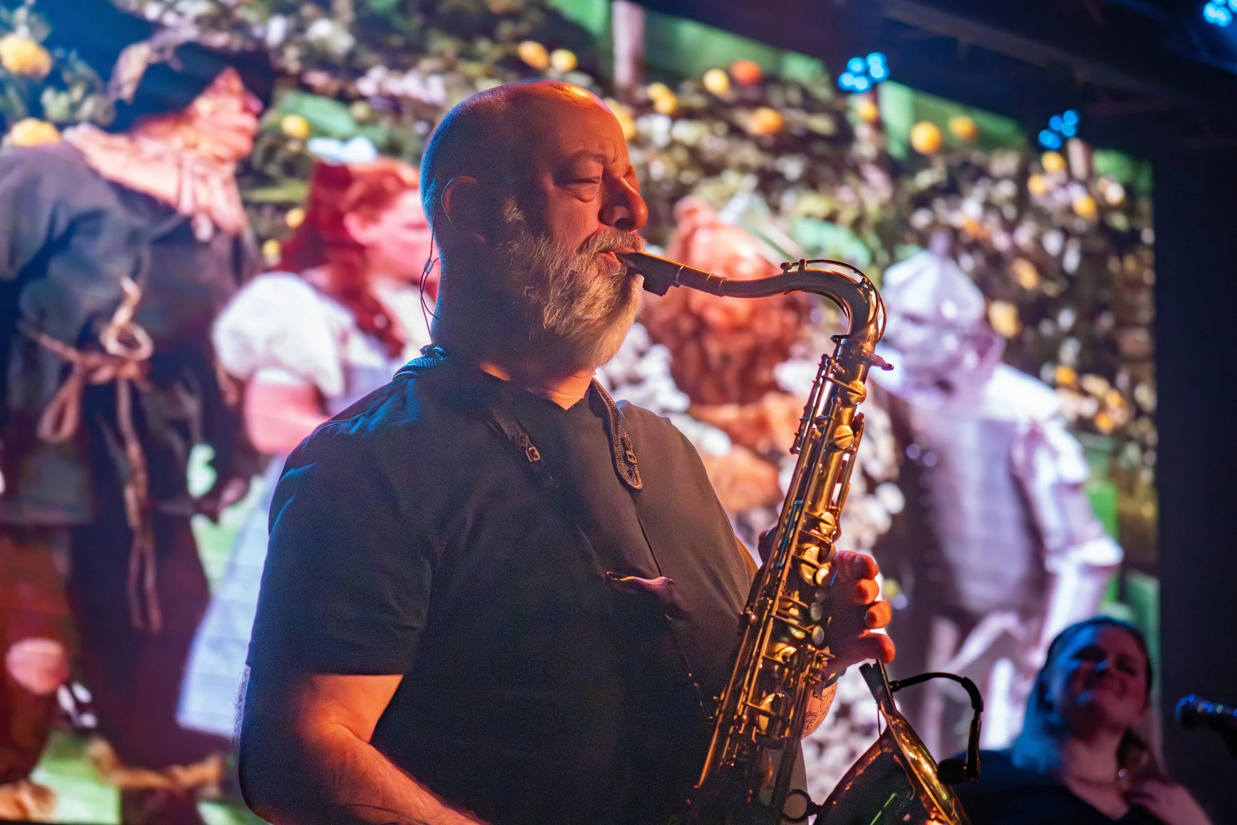 A man with a beard playing a saxophone on stage with a colorful digital backdrop.