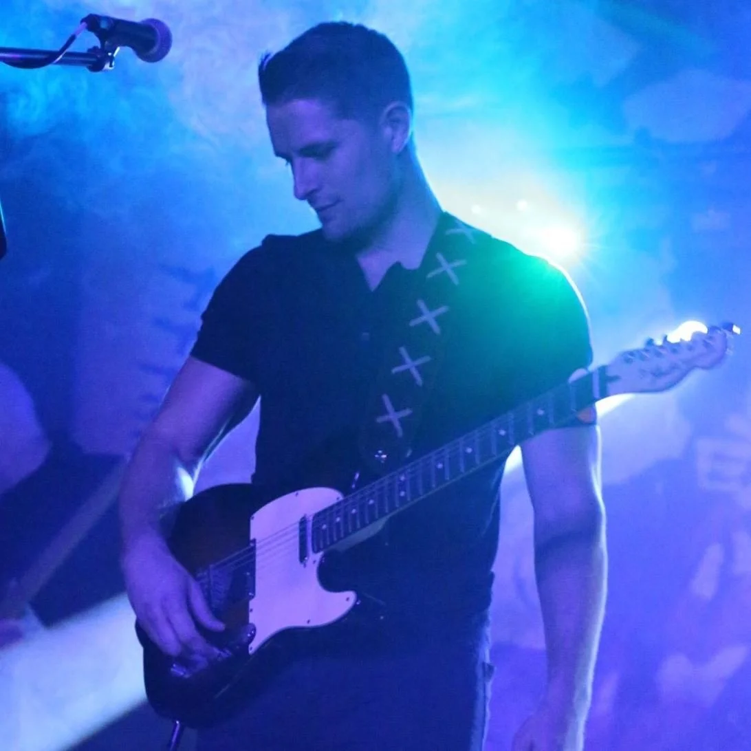 A man playing an electric guitar on stage, illuminated by blue and green stage lighting, with a microphone overhead.