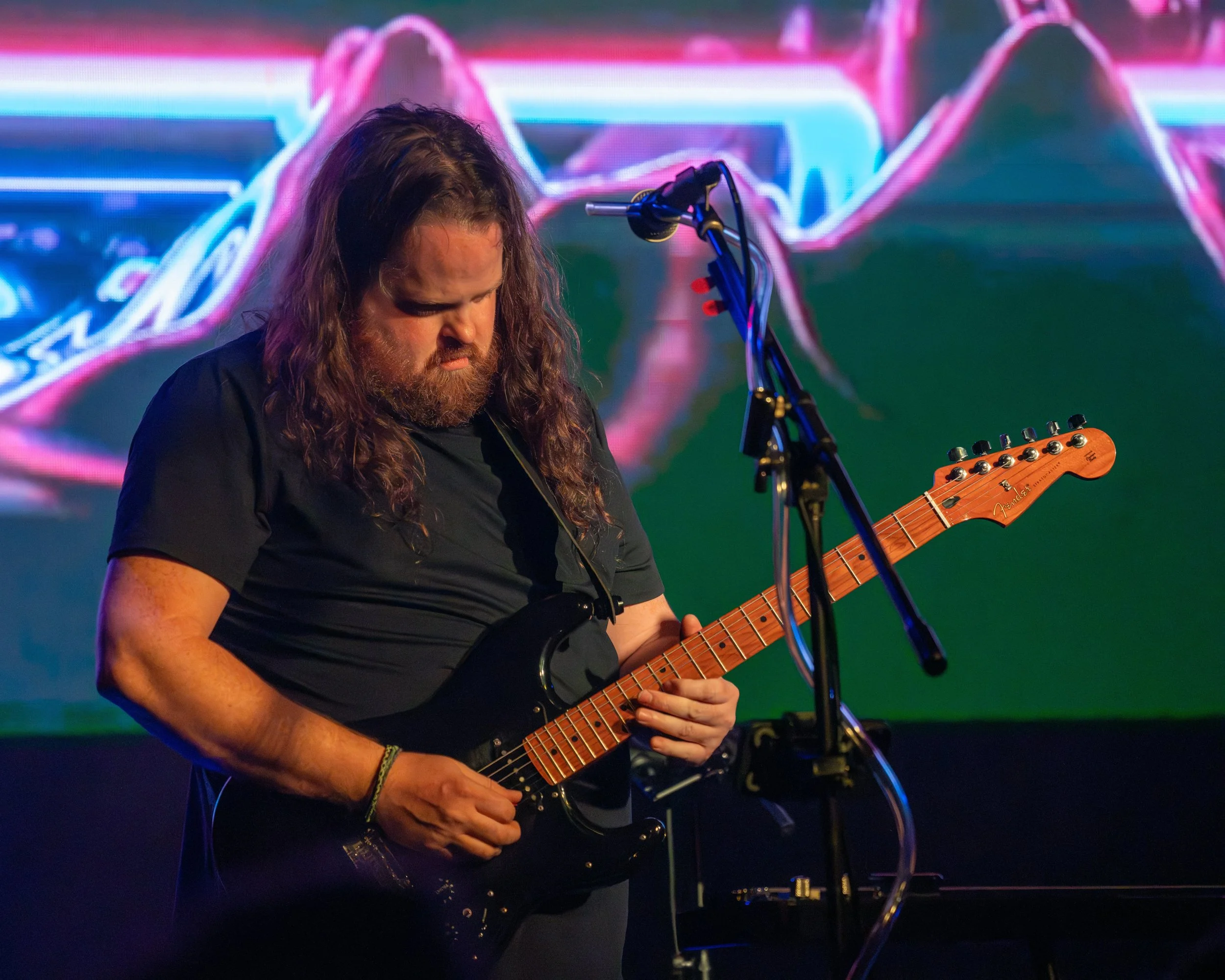 A man with long curly hair and a beard playing an electric guitar on stage, with a colorful digital background behind him.