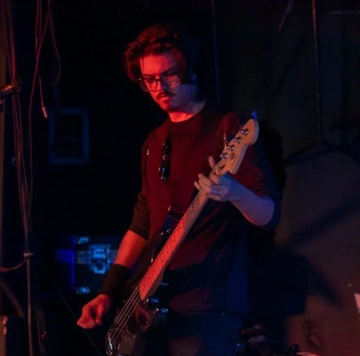 A man playing an electric bass guitar on stage during a live music performance, with drums and microphones in the background.