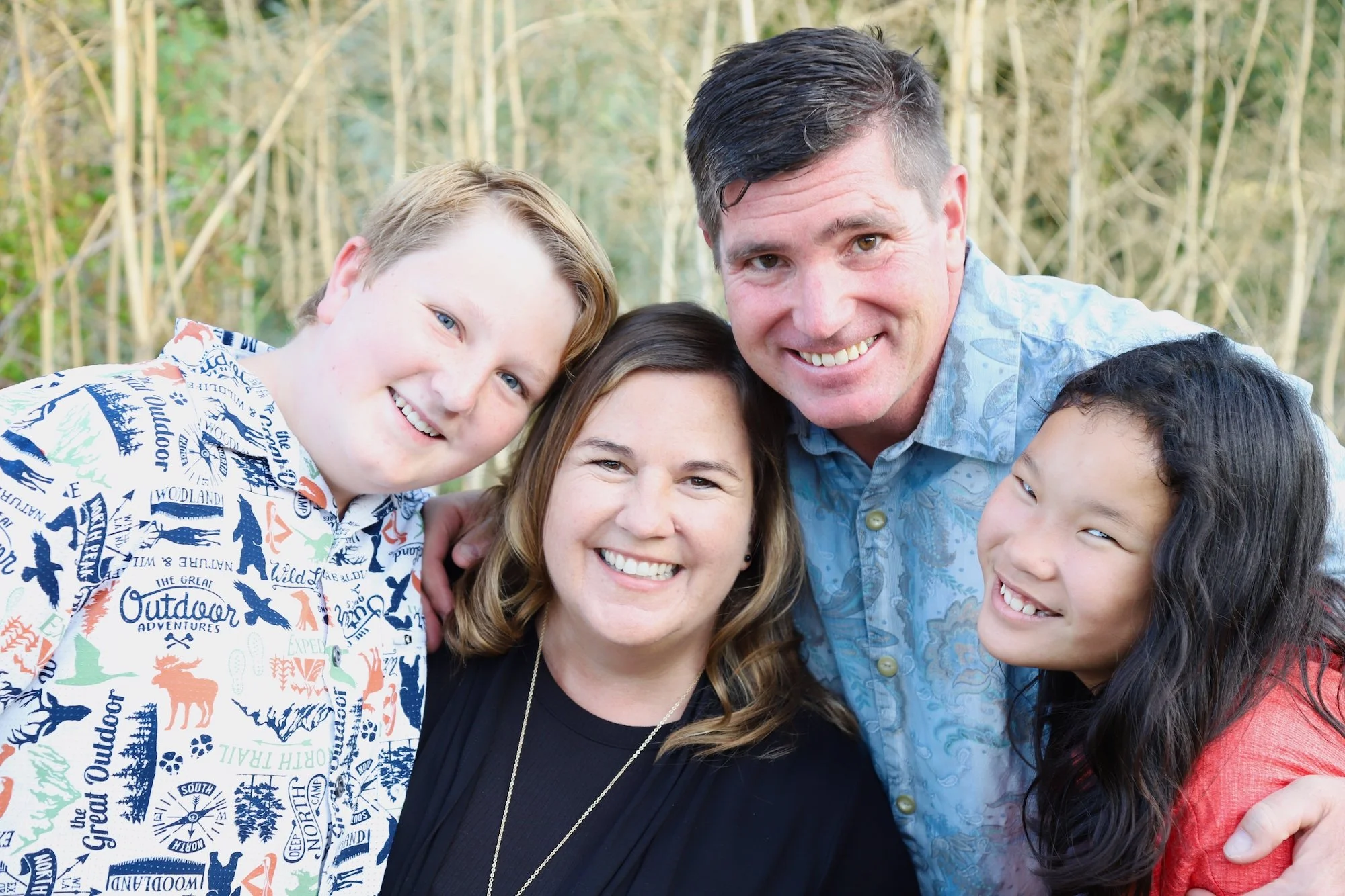 A happy family of five smiling together outdoors with trees in the background.