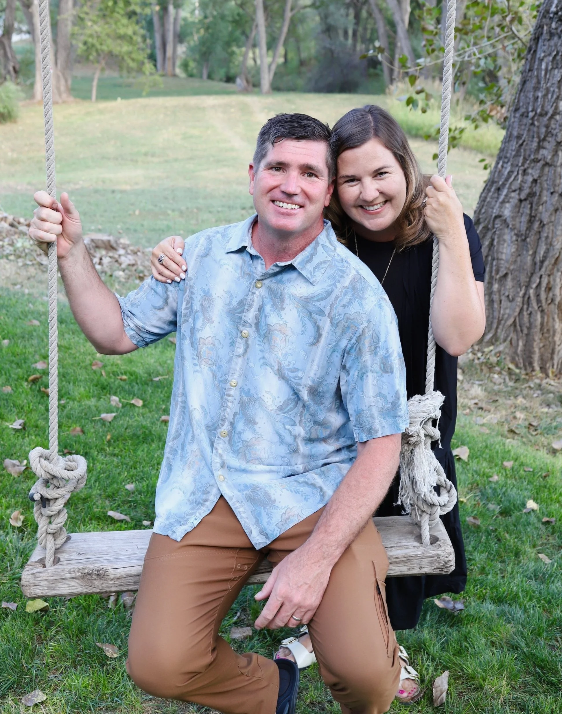 A couple sitting on a wooden swing in a park with green grass and trees, smiling at the camera.