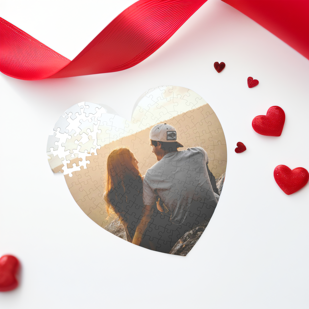 A heart-shaped jigsaw puzzle featuring a photo of a couple sitting outdoors at sunset, with red hearts and a red ribbon around the puzzle.
