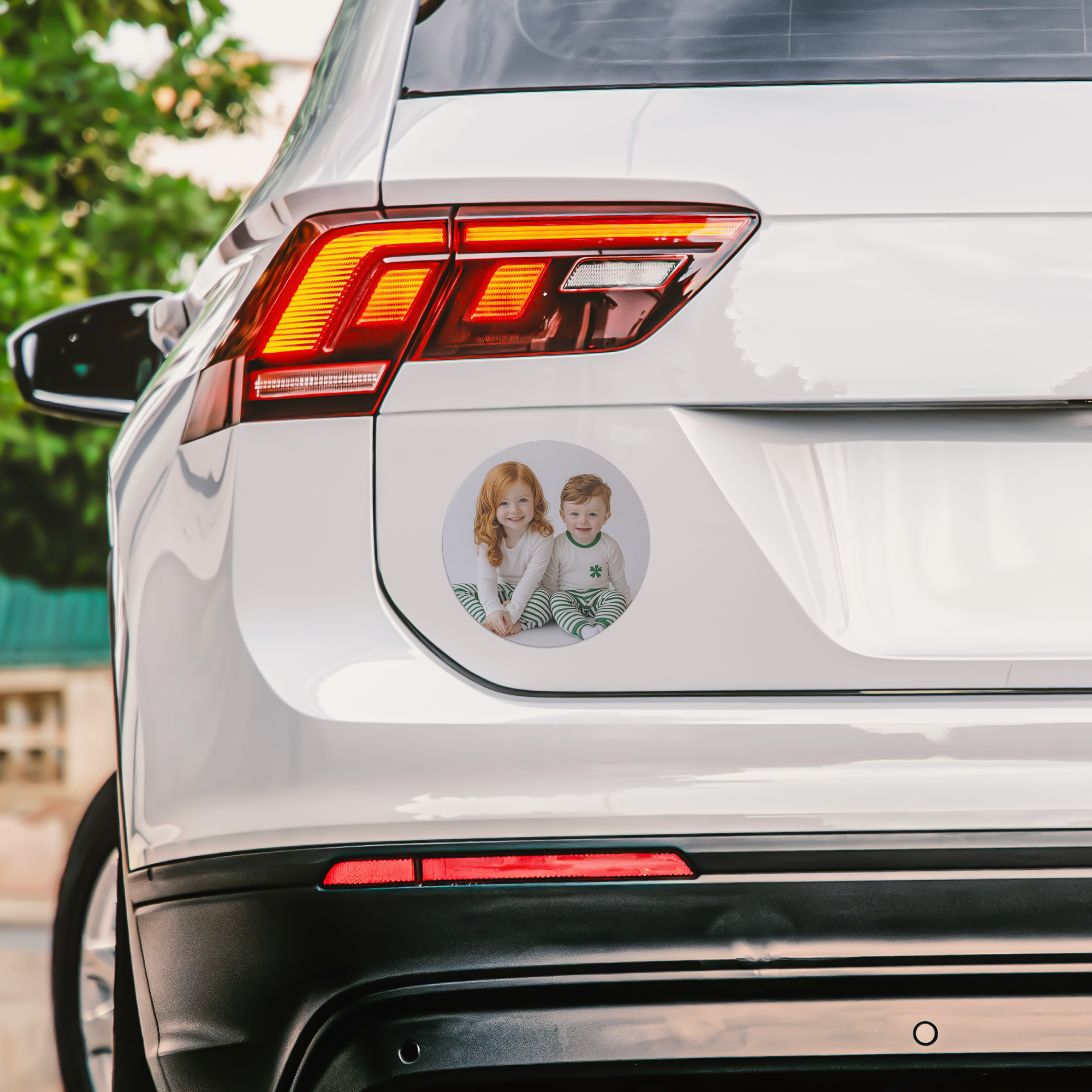 Close-up of a white car's rear with a round sticker of two children sitting together on the trunk. The children are a girl with red hair and a boy with brown hair, both wearing pajamas with horizontal striped pants.
