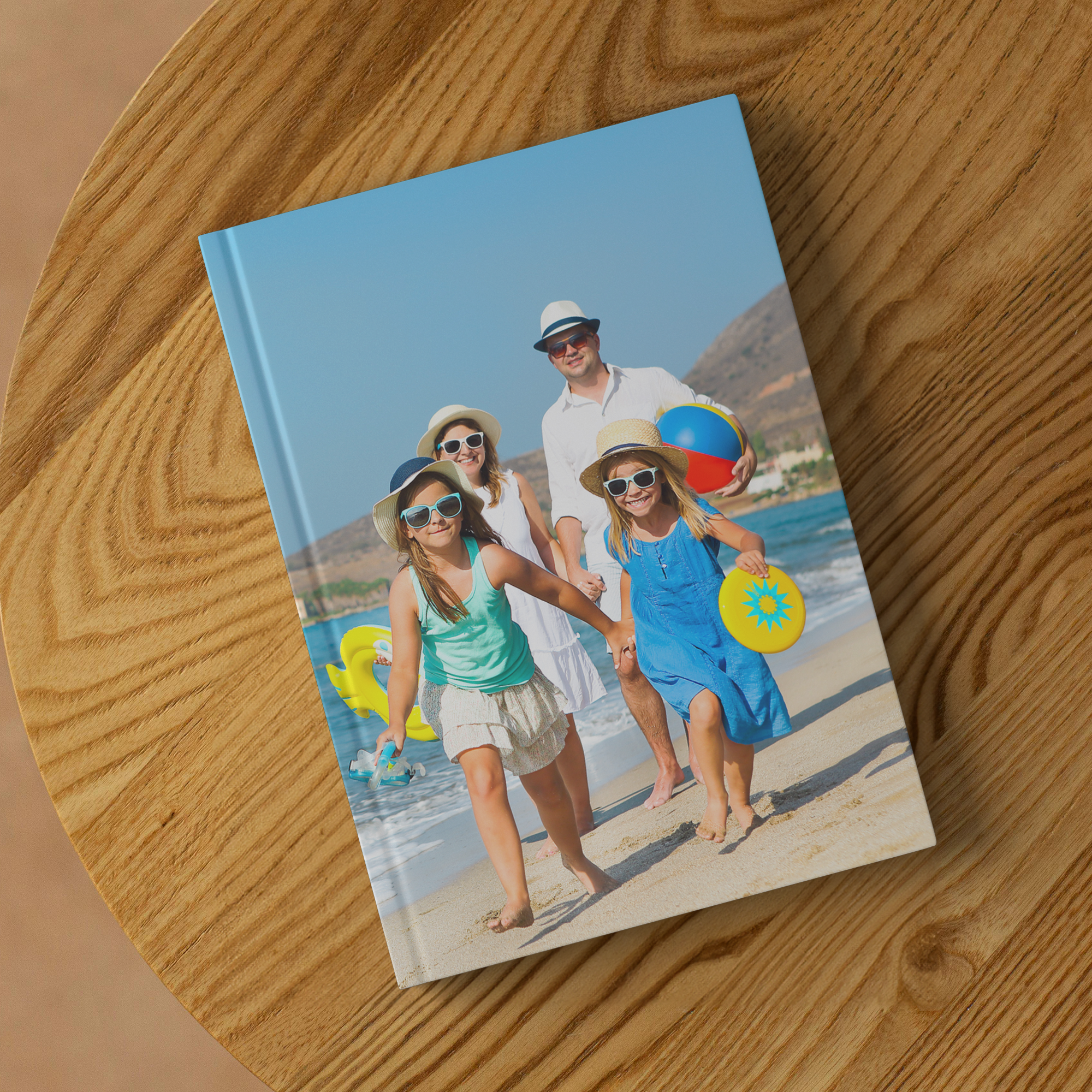 A group of five people, including three women and two men, walking on a beach in summer. They are wearing swimsuits, sunglasses, and sun hats, and are holding beach balls and toys. The background shows a seaside with hills and a clear sky. The photo 