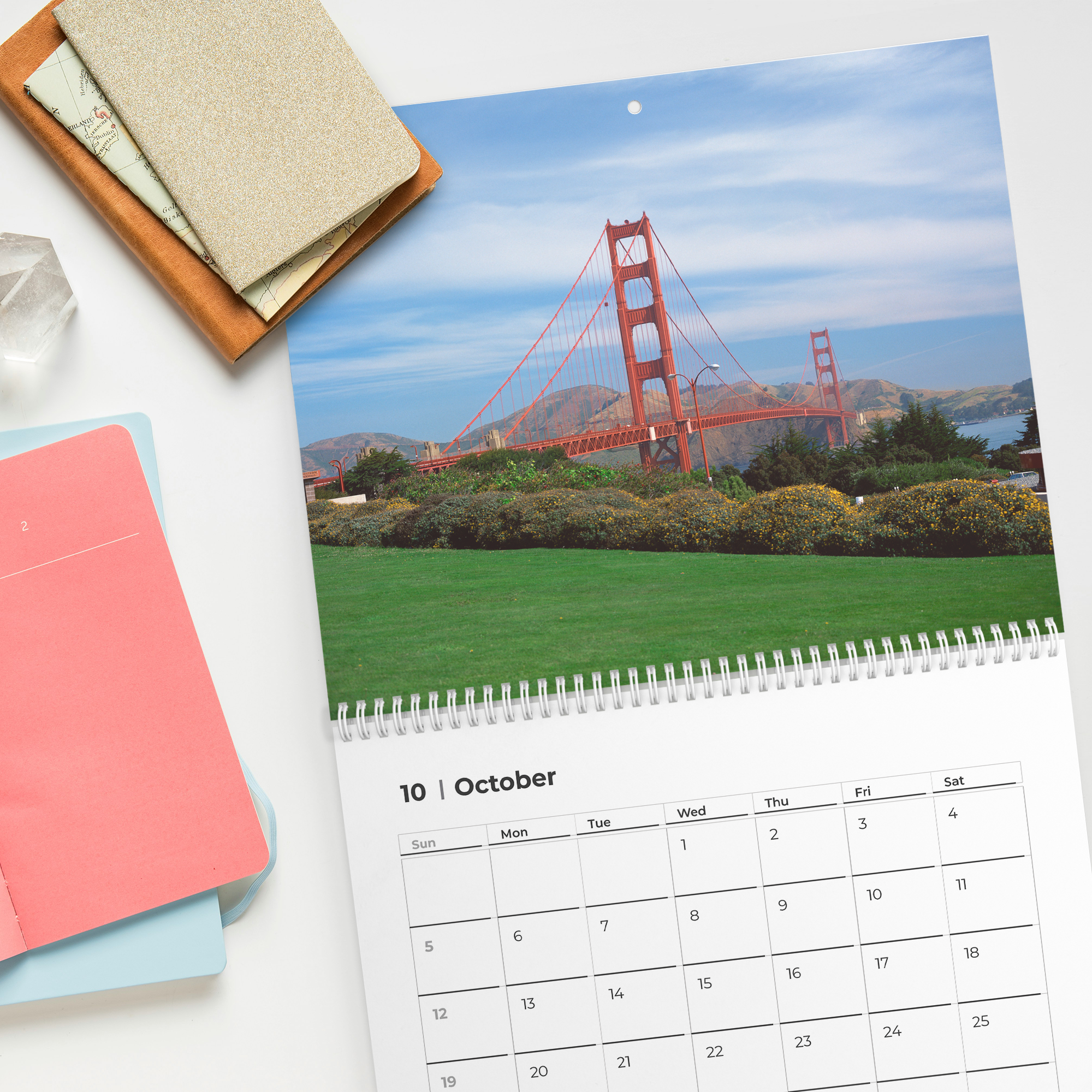 A desk calendar displaying October 10th, a photo of the Golden Gate Bridge in San Francisco, a pink notepad, a blue notepad, a notebook with a textured cover, a map, and a crystal on a white surface.