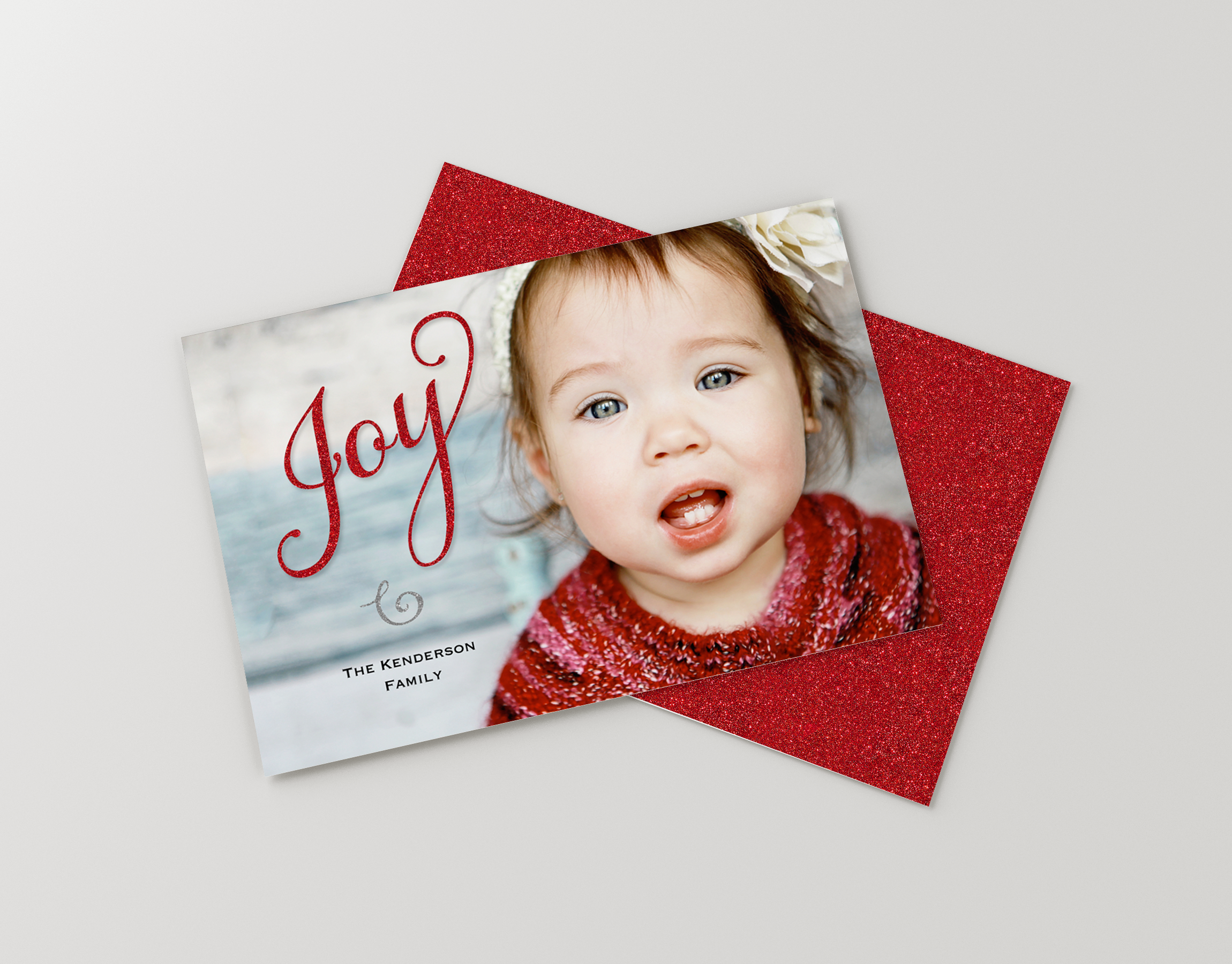 A holiday greeting card featuring a close-up of a young girl with blue eyes and brown hair, wearing a red sweater with white snowflakes. The card has red glittery borders and the words "Joy" and "Family" visible. The card is placed on top of a red gl