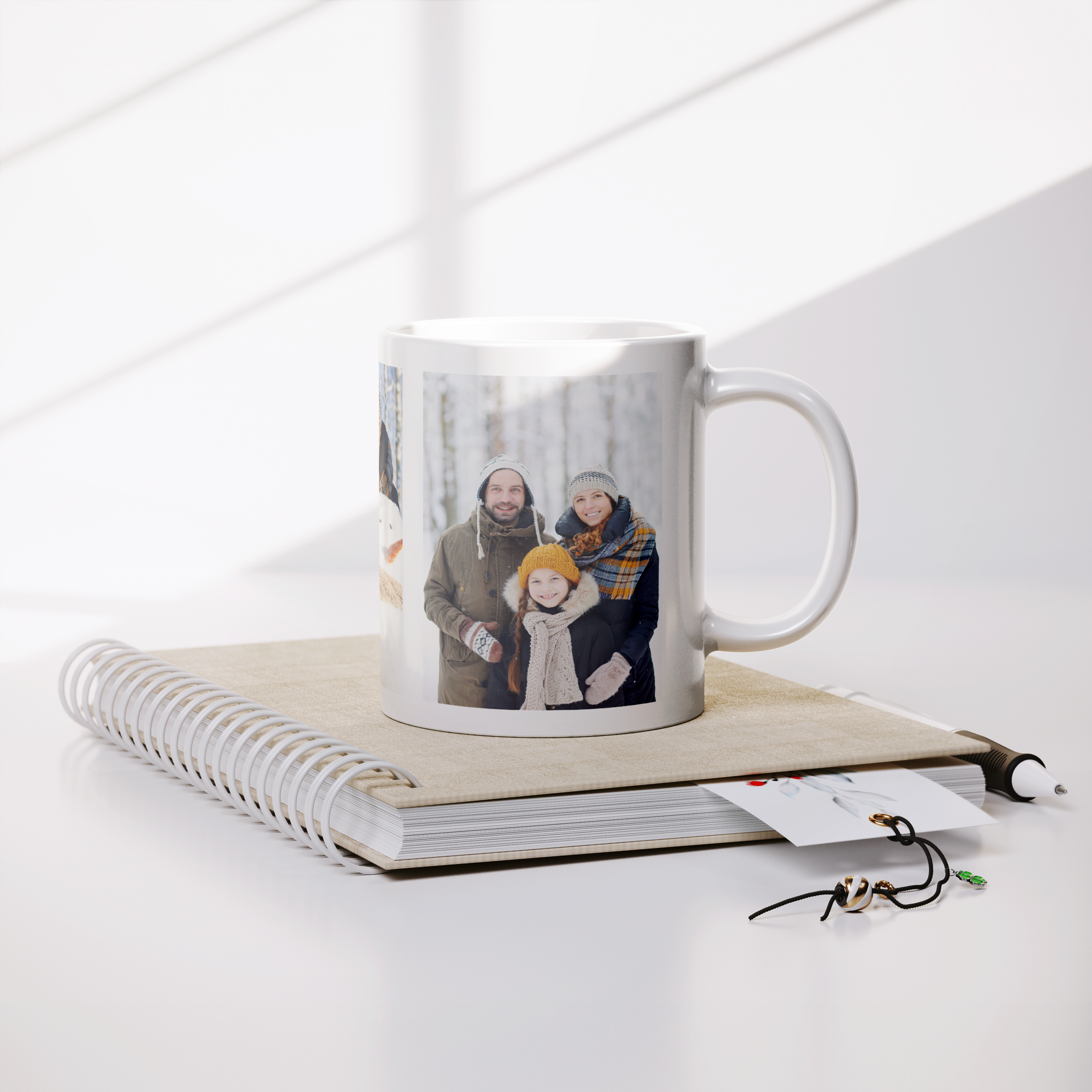 Close-up of a white ceramic mug with a printed photo of a happy family outdoors in winter, surrounded by snow, wrapped in warm clothing and knit hats, placed on top of a notebook with coiled binding and some jewelry on a white surface with space arou