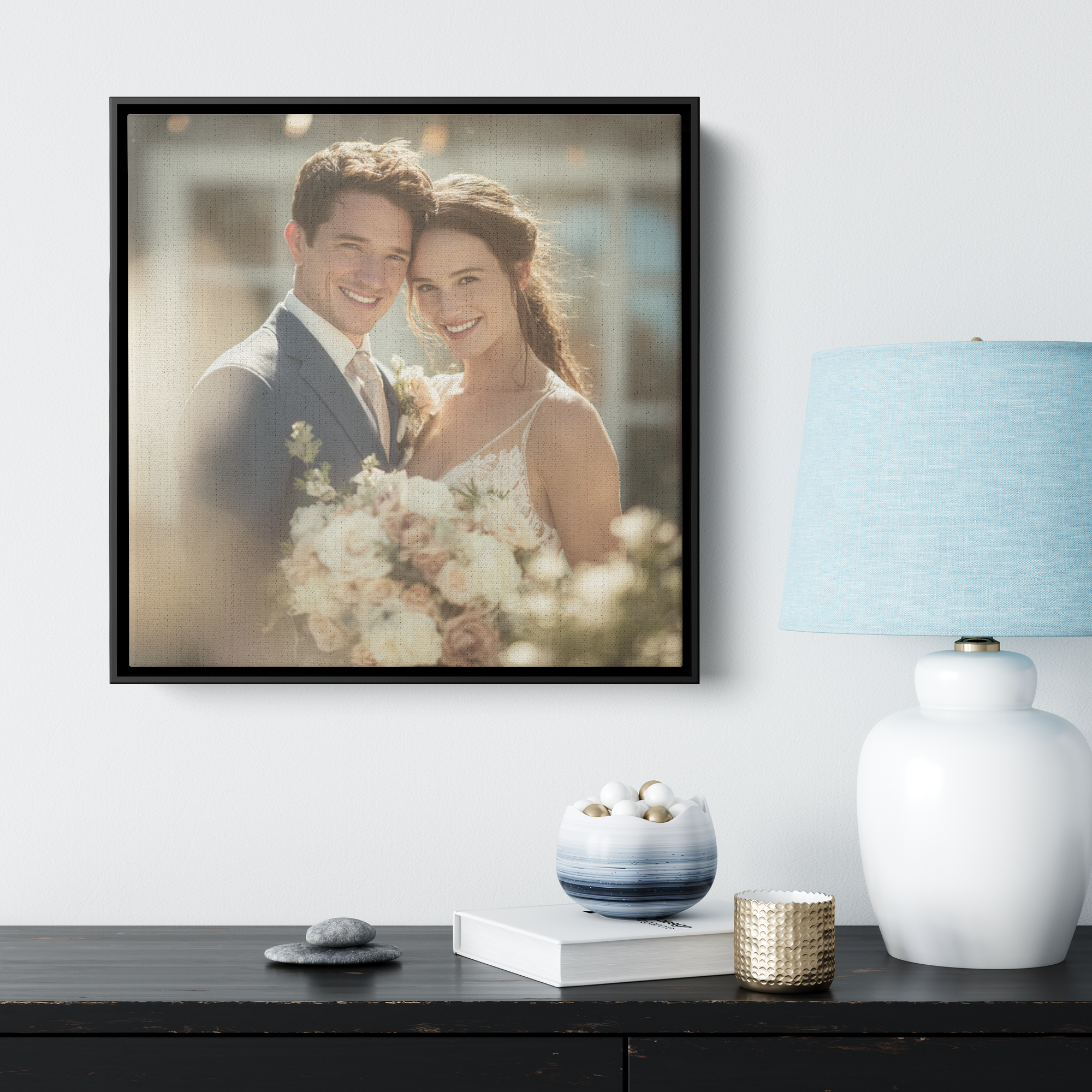 A modern interior decor scene with a framed wedding photo of a smiling couple, a white table with a striped blue and white vase filled with white and blush flowers, a white vase, a smaller textured gold container, and a stack of books, all arranged on a dark wooden surface against a white wall.