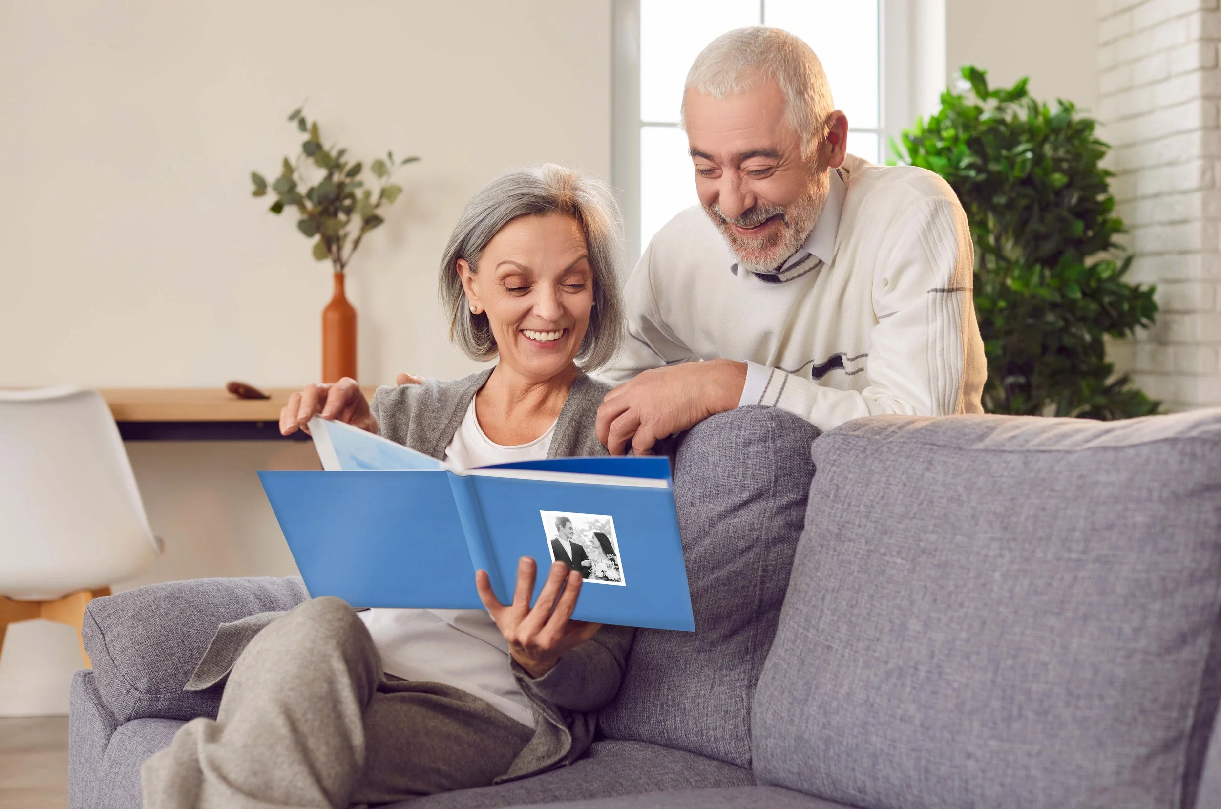 An elderly couple sitting on a gray sofa, looking at a photo album, smiling and sharing a happy moment together in a bright living room with green plants and natural light.