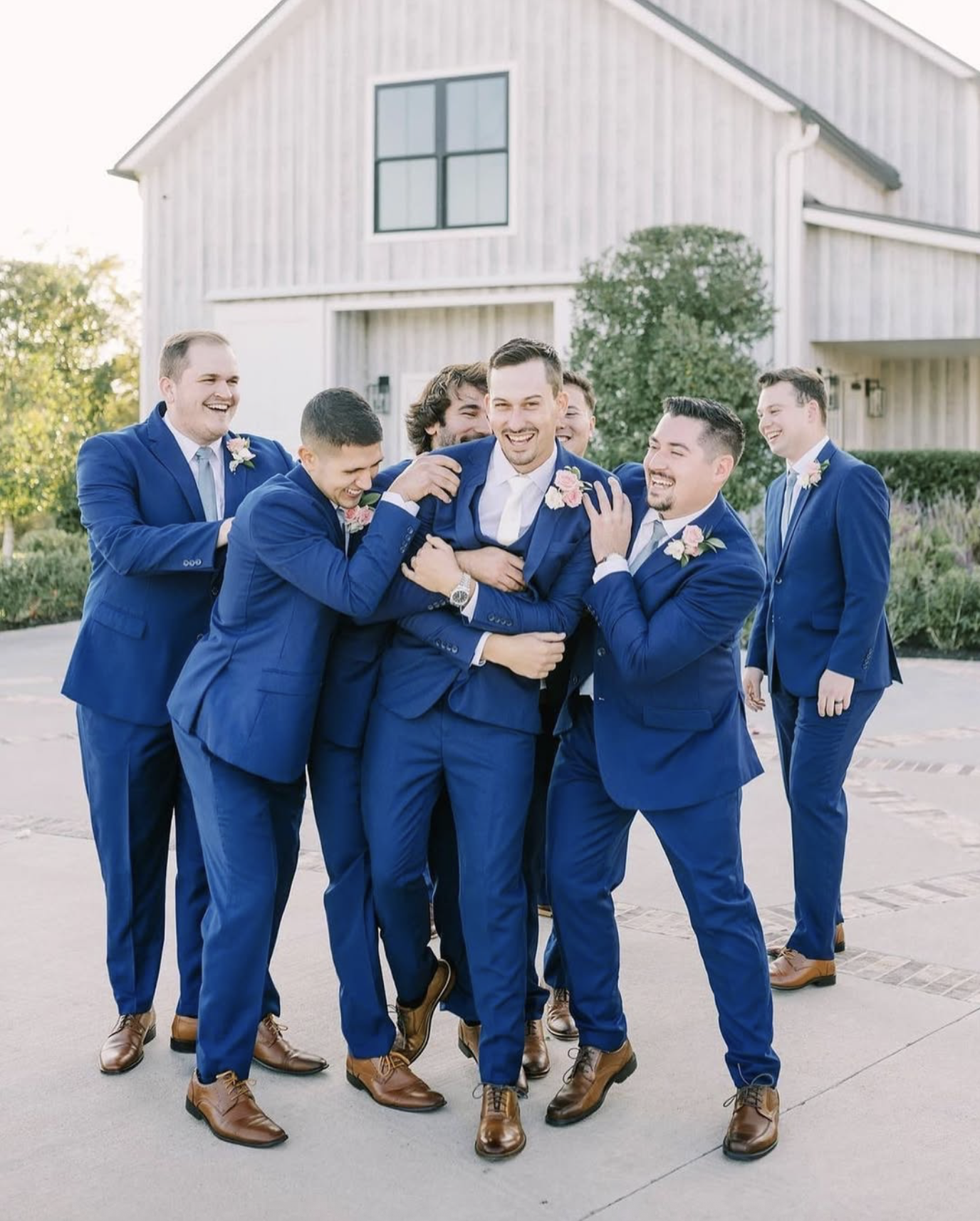A group of seven men in matching blue suits laughing and joking outdoors in front of a modern, light-colored building with greenery.