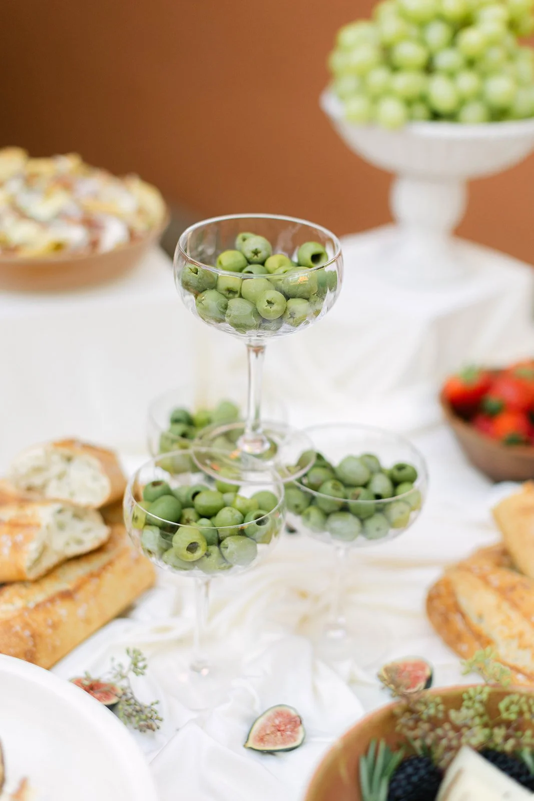 Martini glasses filled with green olives on a draped white table cloth with french bread and figs.