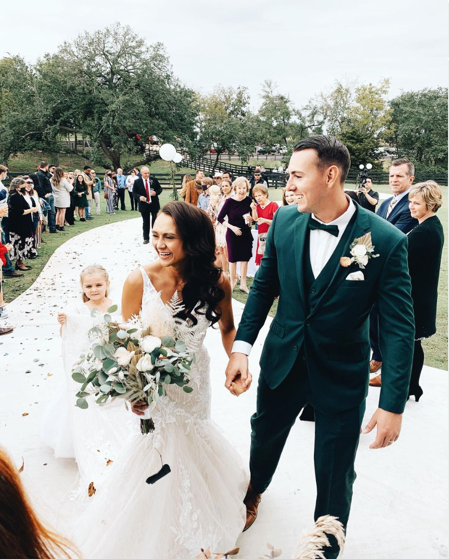 A bride and groom holding hands and smiling during their outdoor wedding ceremony, with guests in the background.