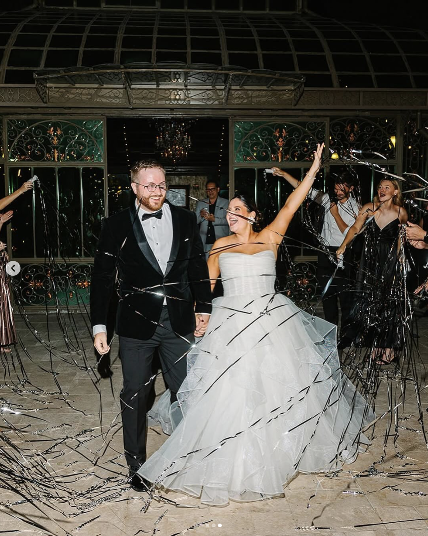 A newlywed couple dancing at their wedding reception, surrounded by guests, with black and silver streamers falling around them in a decorated indoor venue.