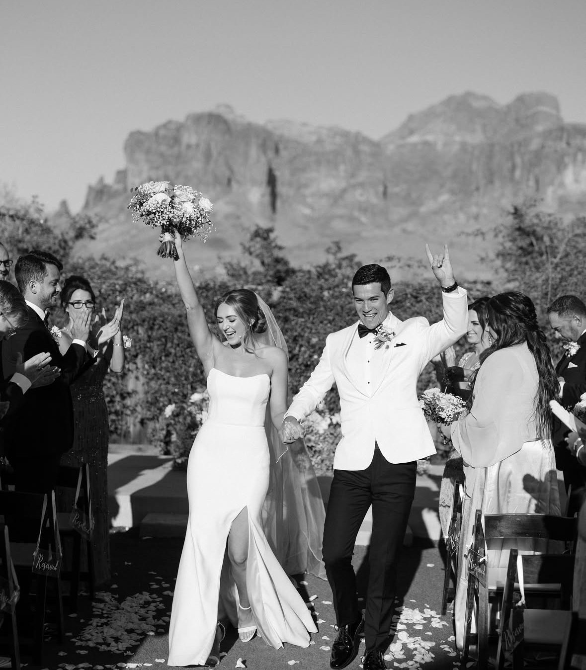 Black and white photo of a wedding celebration with bride and groom walking down the aisle, guests smiling and clapping, outdoor mountain landscape in the background.