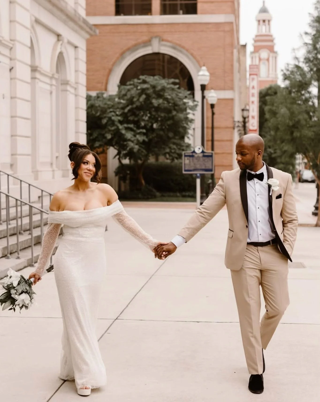A bride and groom holding hands and walking outside on their wedding day, with the bride in a white off-the-shoulder dress holding a bouquet, and the groom in a beige tuxedo with black trim.
