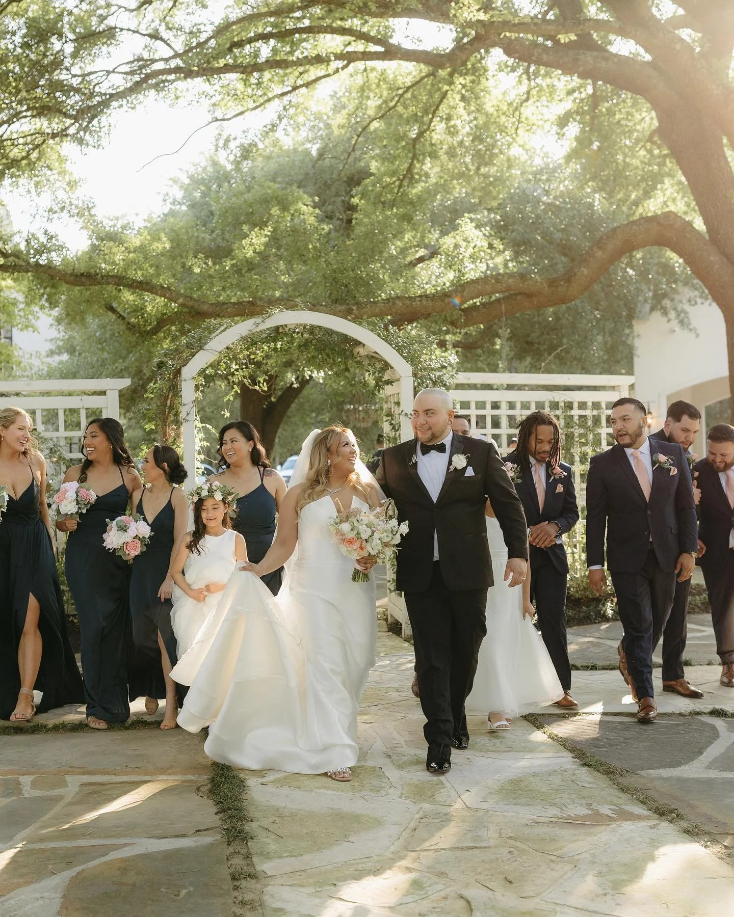 A wedding party walking outdoors under a large tree, with the bride and groom in the center and the bride holding a bouquet, surrounded by bridesmaids and groomsmen.