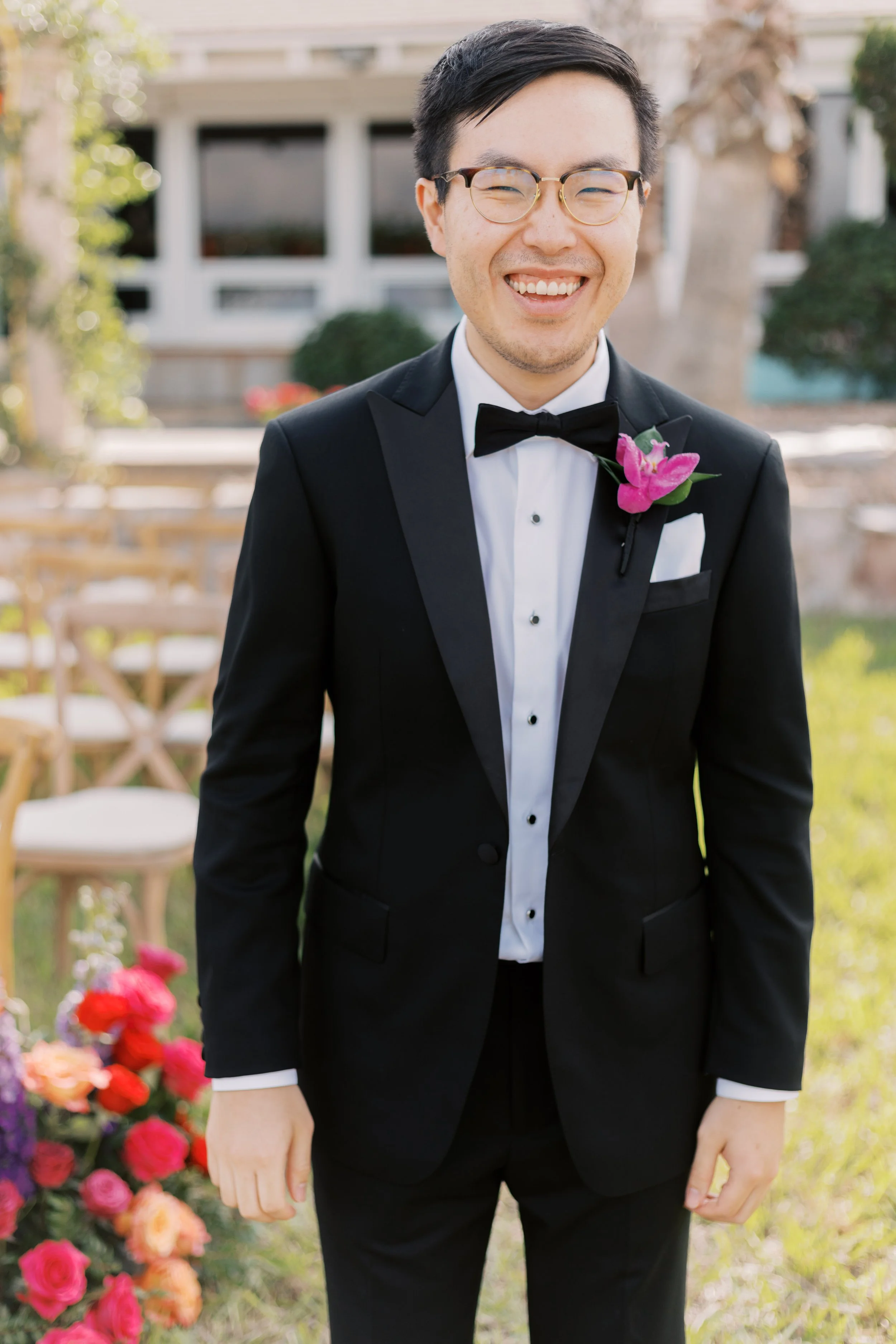 A man wearing a black tuxedo, white dress shirt, black bow tie, and glasses, smiling outdoors with chairs and colorful flowers in the background.
