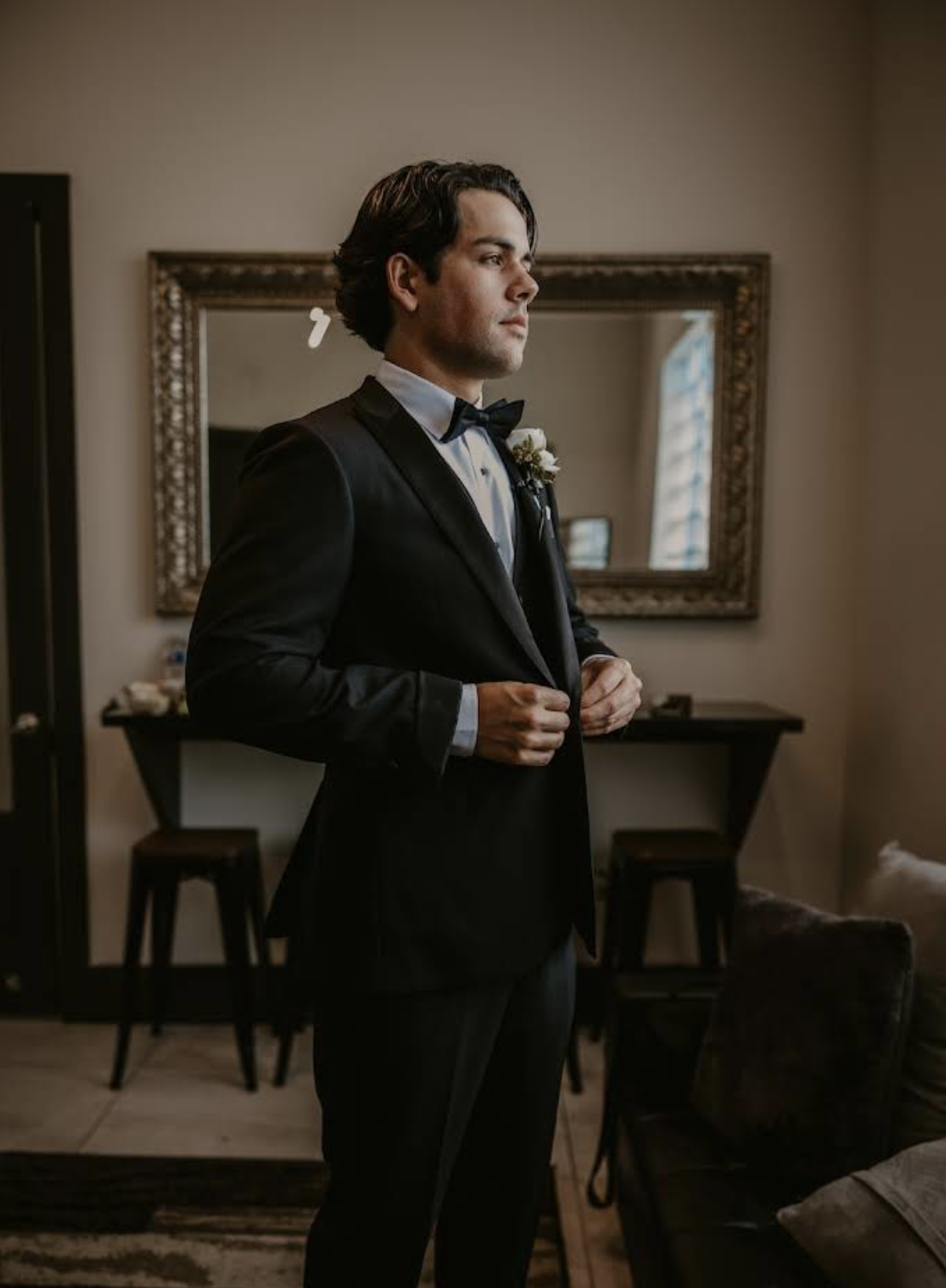 A man in a tuxedo with a bow tie, adjusting his jacket, standing in a room with a mirror and a window in the background.