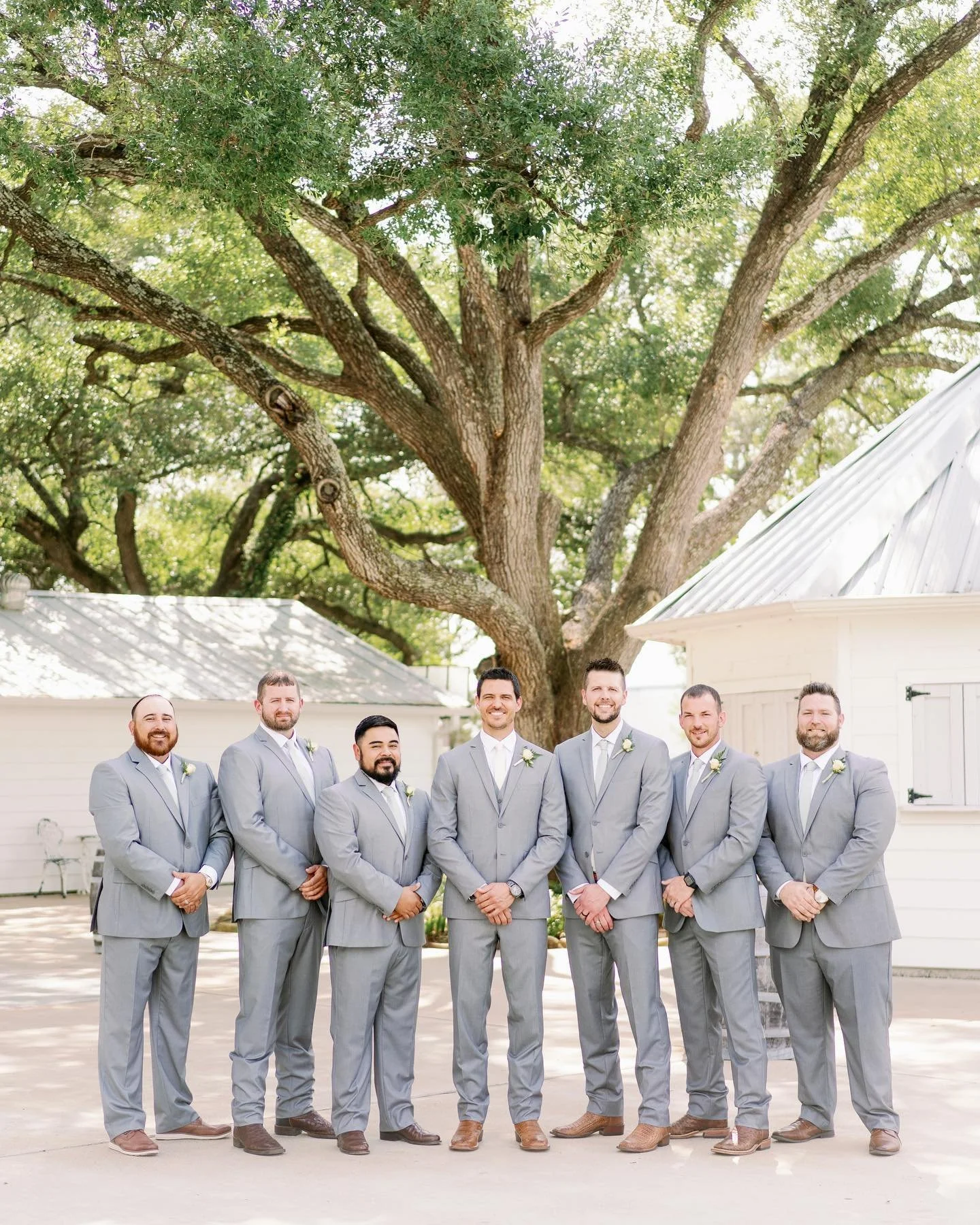 A group of seven men dressed in light gray suits with white shirts and light gray ties, standing outdoors in front of a large tree and white buildings, at a wedding or formal event.